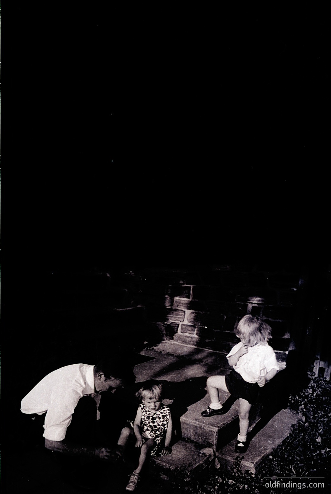 Black-and-white photograph of three children exploring a cemetery at dusk. Adult kneels beside a child in floral dress, while another child sits atop a weathered tombstone. Grave markers and overgrown foliage frame the scene. Likely mid-20th century, urban setting.