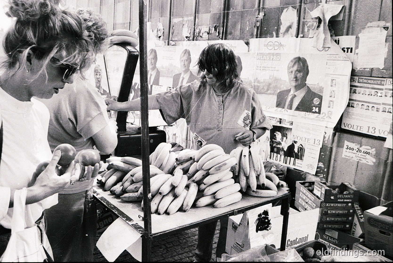 Street vendor stall showcasing bundled sausages and political campaign posters. Two women engage with products—one handling sausages, the other examining a poster. Posters feature candidates with text in Cyrillic, suggesting Eastern Bloc region. Mid-20th century (1960s–1980s) election atmosphere.