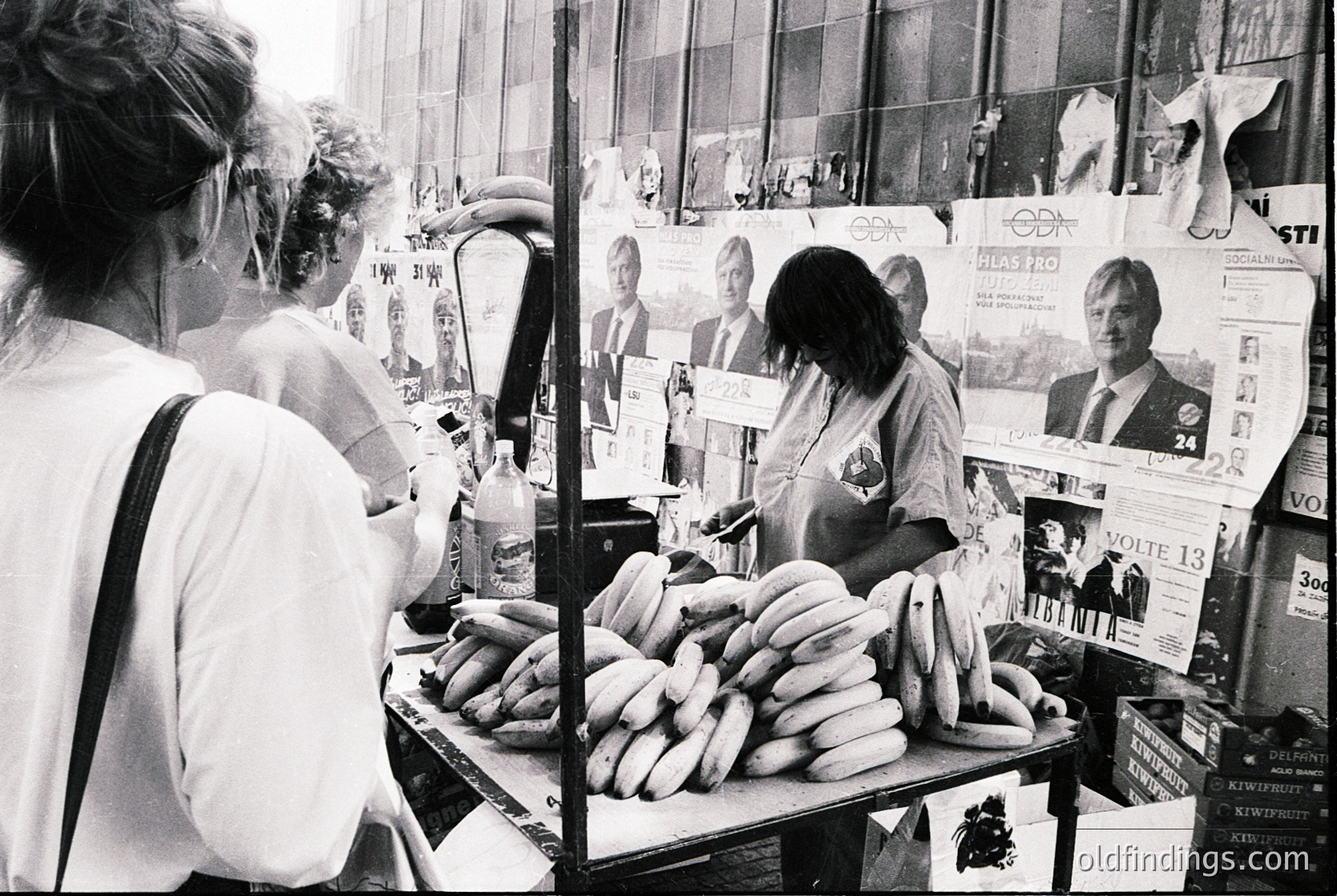 Street vendor stall displaying sausages in a 1970s Eastern Bloc market. Posters of political figures and election candidates adorn the backdrop. Women in casual attire examine the goods.