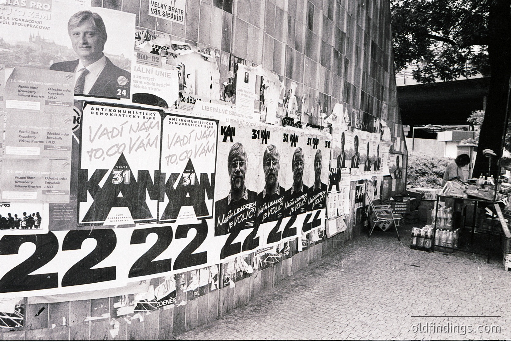 Wall covered in anti-communist campaign posters, featuring prominent figures and slogans like "KAN KAN" and "22.22" alongside handwritten text in Czech. Political rally or protest setting, likely late 20th century Eastern Bloc.