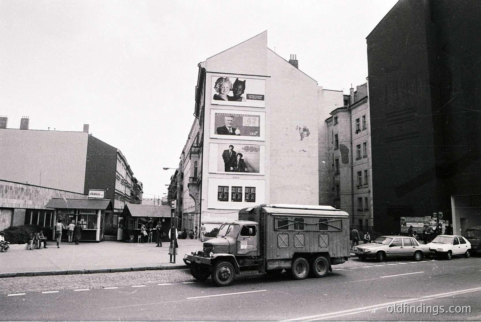 Urban street scene featuring a mid-20th-century European cityscape. Prominent white building with large posters of a cat and formal portraits, likely from the 1960s–70s. Truck with "X" markings parked on cobblestone road; surrounding older architecture and parked cars.