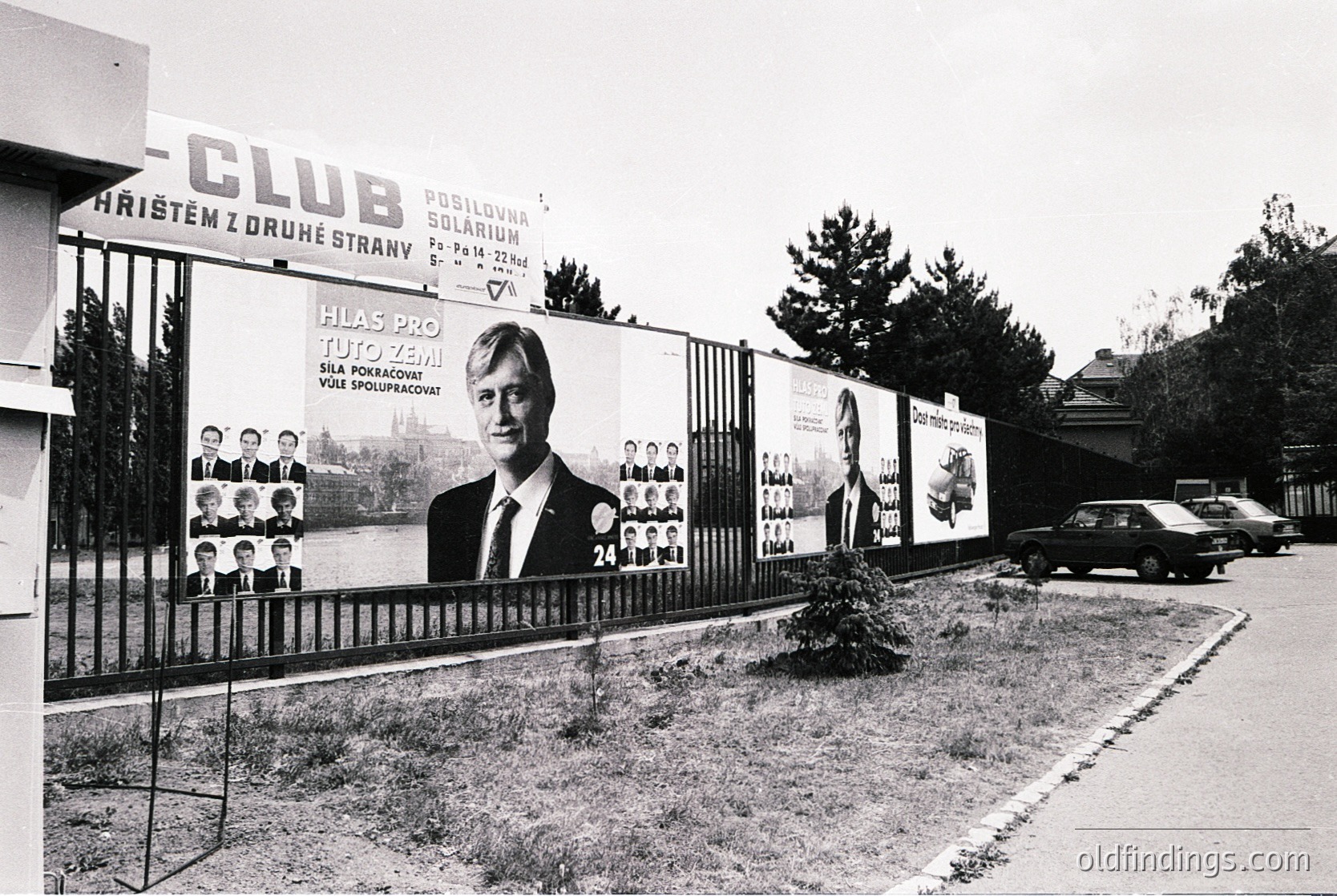 Black-and-white street scene featuring a **1970s-era political campaign billboard** for a candidate named "Tuto Zemana" in Czechoslovakia. The billboard, mounted on a chain-link fence, displays his portrait alongside smaller images of supporters. Adjacent signage promotes a "Club" with solarium services. A vintage car and residential buildings appear in the background.