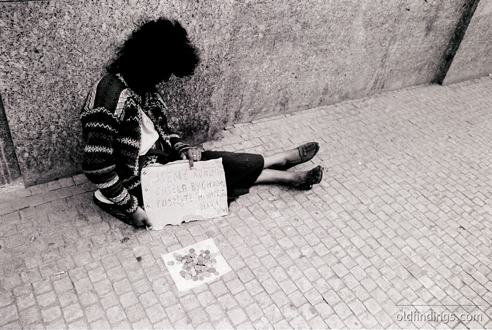 A person sits on a cobblestone street, leaning against a concrete wall, holding a handwritten sign reading *"I am a woman, help me, please. I have a child."* Coins and a small cardboard tray lie beside them. Black-and-white urban street scene, likely 1970s–1990s.