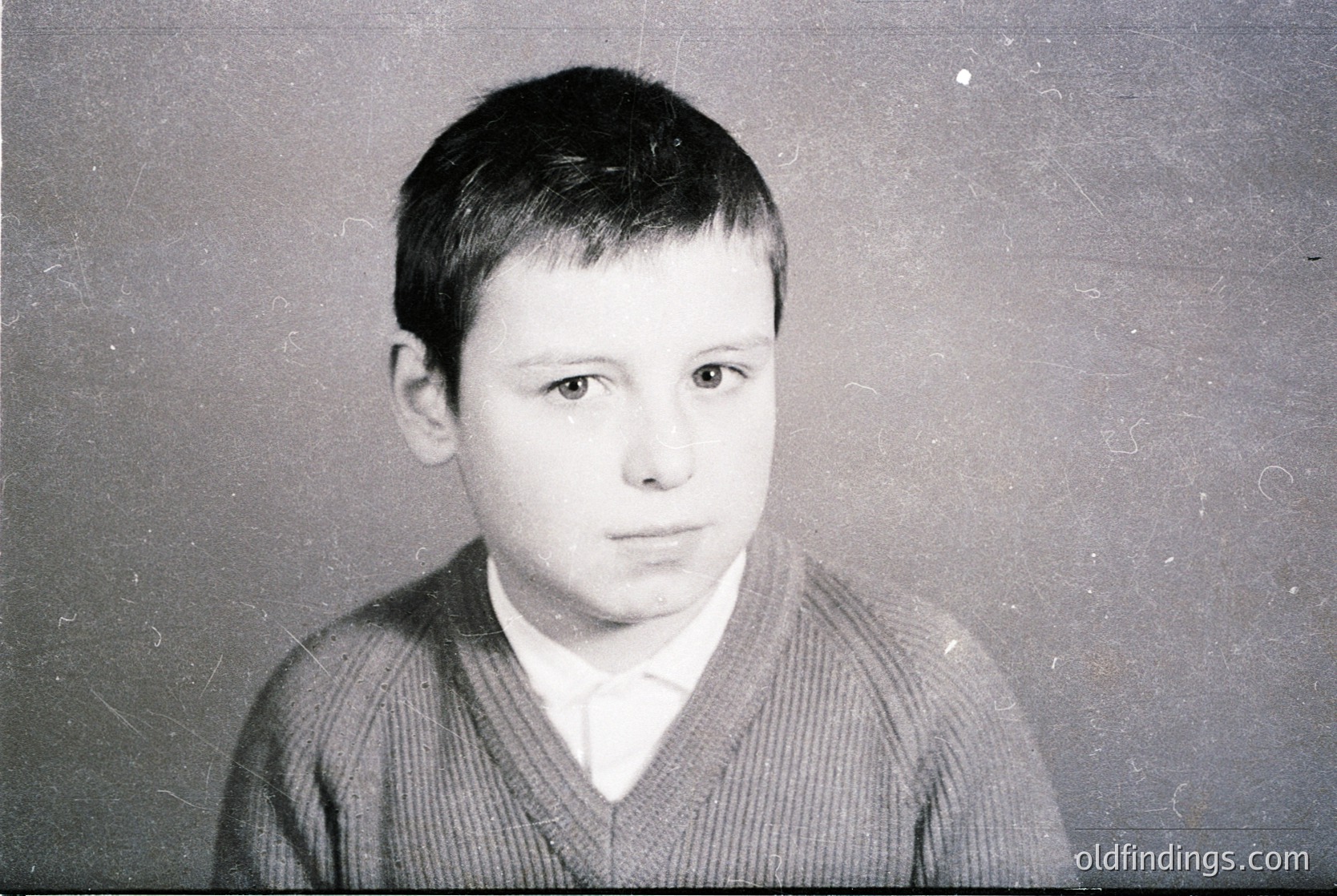 Portrait of a young boy in a vintage-style black-and-white photo, likely from the 1960s–1980s. He wears a V-neck sweater and a white collared shirt, posing against a plain background. Expression is neutral, with short, neatly combed hair.