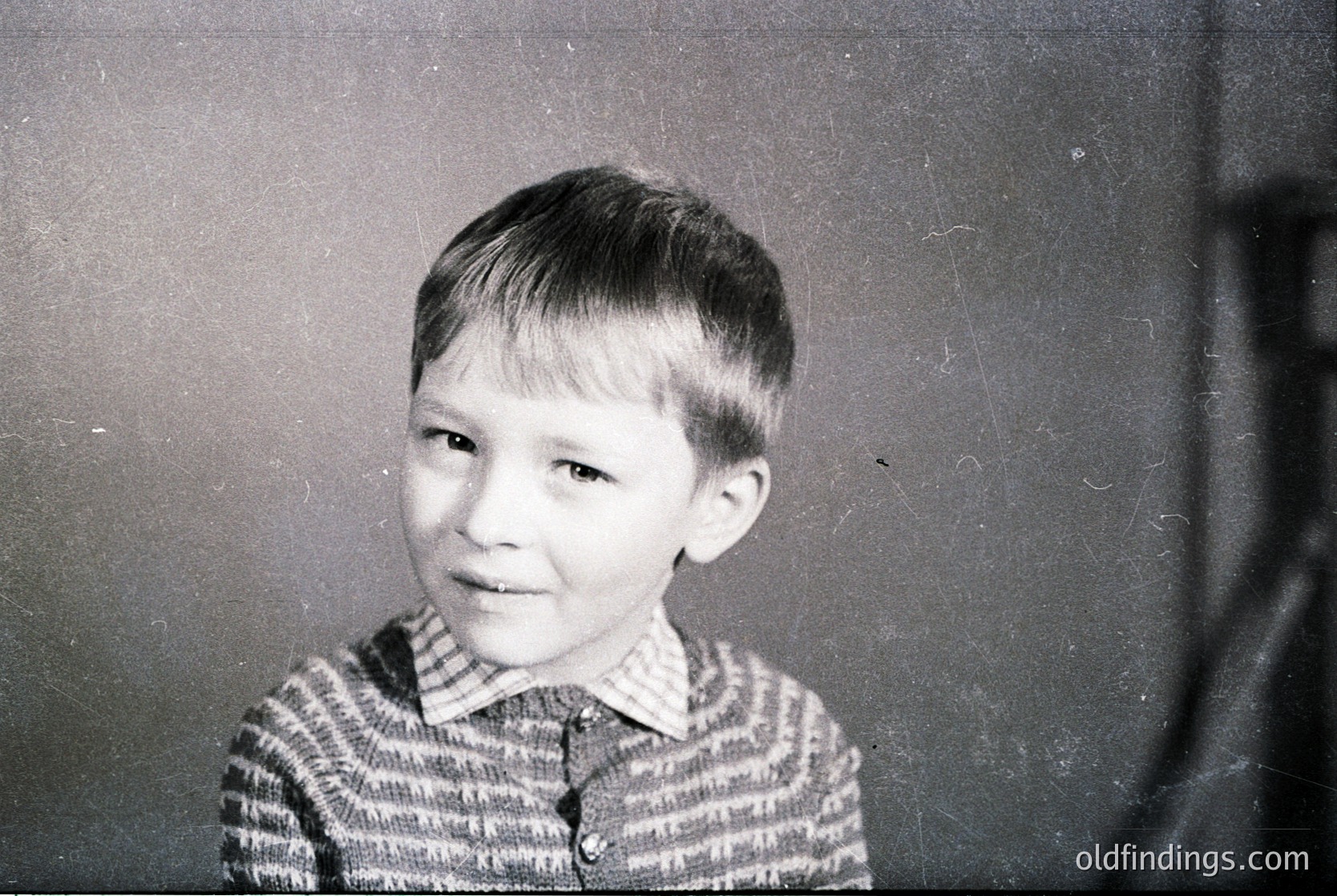Vintage black-and-white portrait of a young boy with short, neatly parted hair, wearing a striped sweater. Indoor setting with plain wall background, suggesting mid-20th century family photography. Authentic candid expression captures innocence and timelessness.