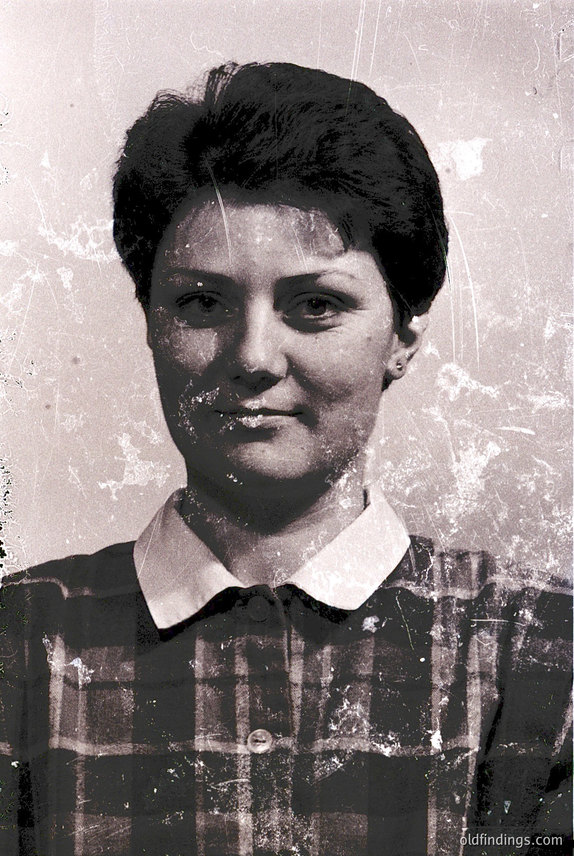 Vintage black-and-white portrait of a woman in a plaid blouse, likely from the 1950s–1970s. Plain background suggests studio or formal setting. Distinctive short, voluminous hairstyle and minimal makeup reflect mid-century fashion trends.