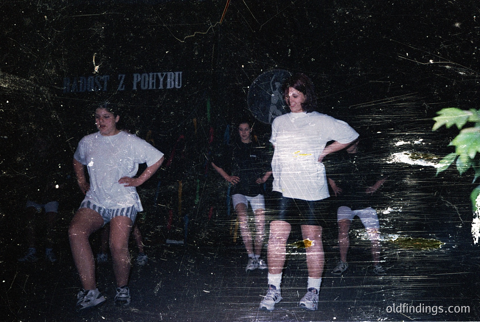 Vintage photo of three individuals posing against a dark, textured wall with Czech signage ("JÁROST Z POBYTU"). Casual 1980s–90s attire: oversized white shirts, rolled-up pants, and sneakers. Blurred reflections and grainy texture suggest analog film. Urban, likely Eastern European setting.