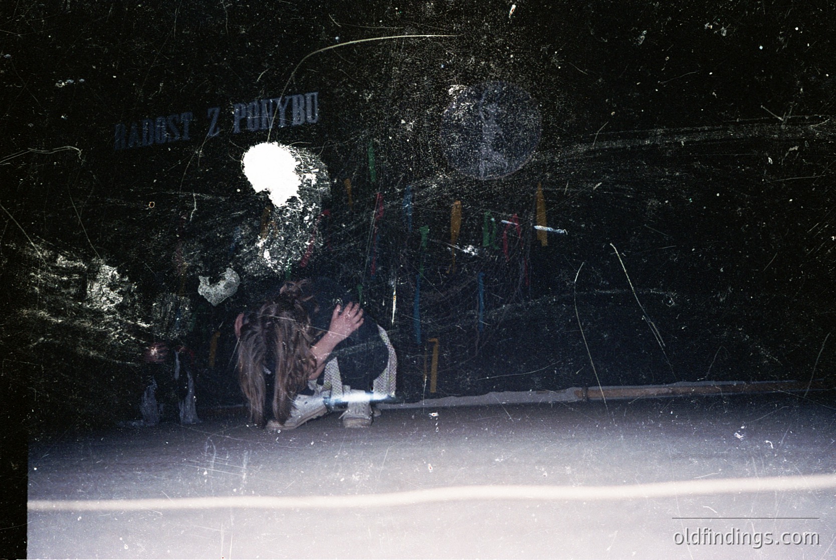 A low-angle shot of a shattered storefront window with Cyrillic signage ("ЗАПОВІД") partially visible. Debris and broken glass litter the scene, revealing a person kneeling inside, possibly clearing glass. Urban decay and post-disaster cleanup evident. Likely Eastern European location, mid-20th century architectural style.