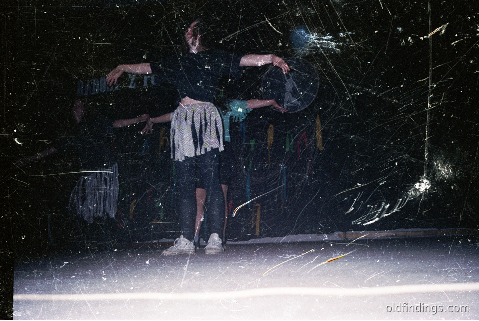 Two dancers in mid-motion against a shattered glass backdrop, likely a stage setting. The performer on left wears a sleeveless top and shorts, while the other in a long skirt and cropped top. Dynamic lighting and glass shards create abstract patterns. Evokes 1990s–2000s contemporary dance or performance art.