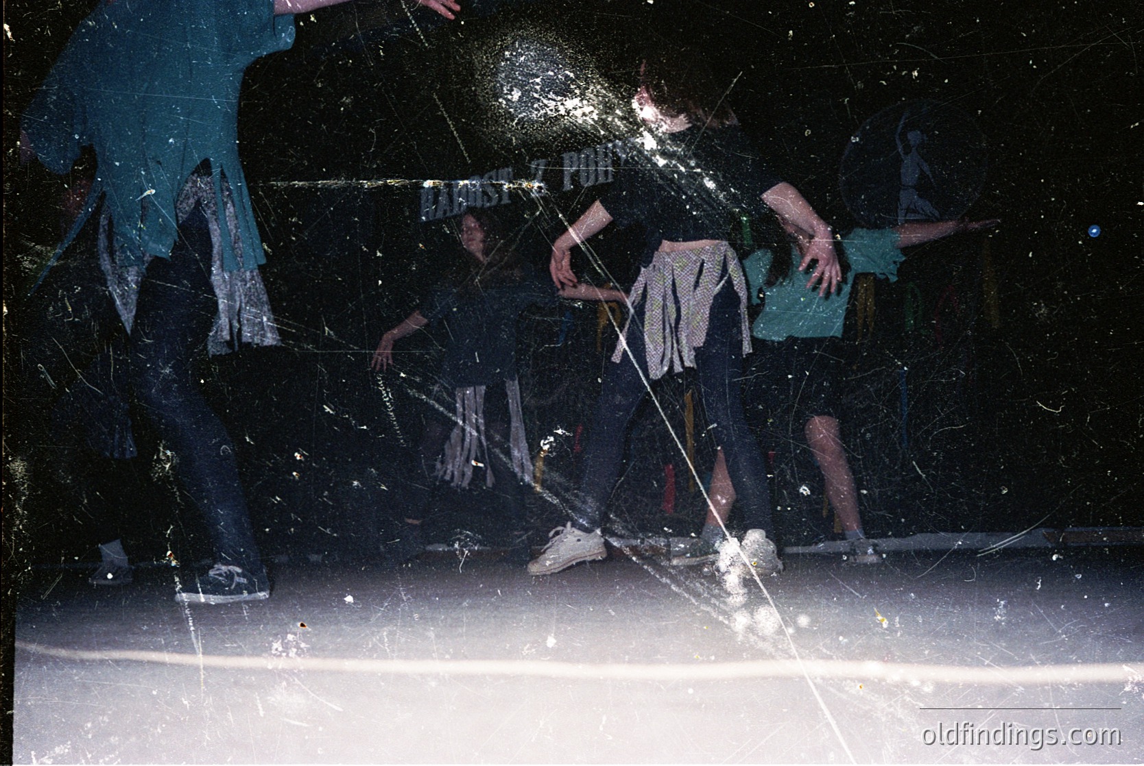 Vintage black-and-white photo capturing an indoor ice hockey game in progress. Players in classic 1970s attire—long-sleeve jerseys, shorts, and knee pads—compete on a rink with visible ice glare. One player in motion, stick raised, while others skate defensively. Reflective lighting and motion blur emphasize dynamic action. Ideal for sports history or retro design references.