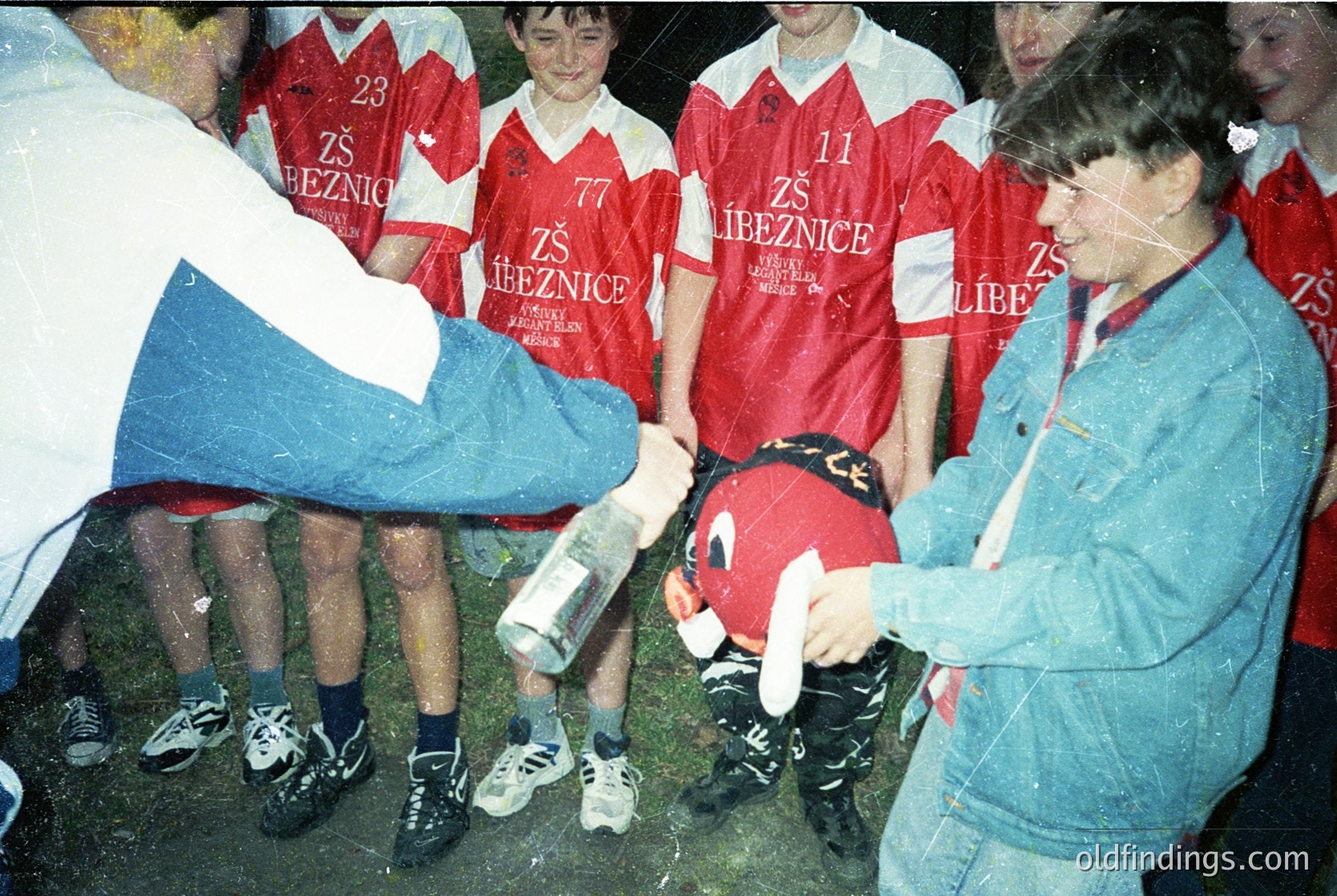 Group huddle with youth soccer team in red jerseys bearing "ZŠ Líbeznice" (likely a Czech school team) and Czech flag scarves, 1980s-1990s. Coach or official hands a red helmet to a player, indicating a goalie drill. Grass field and casual attire suggest amateur or school league play.