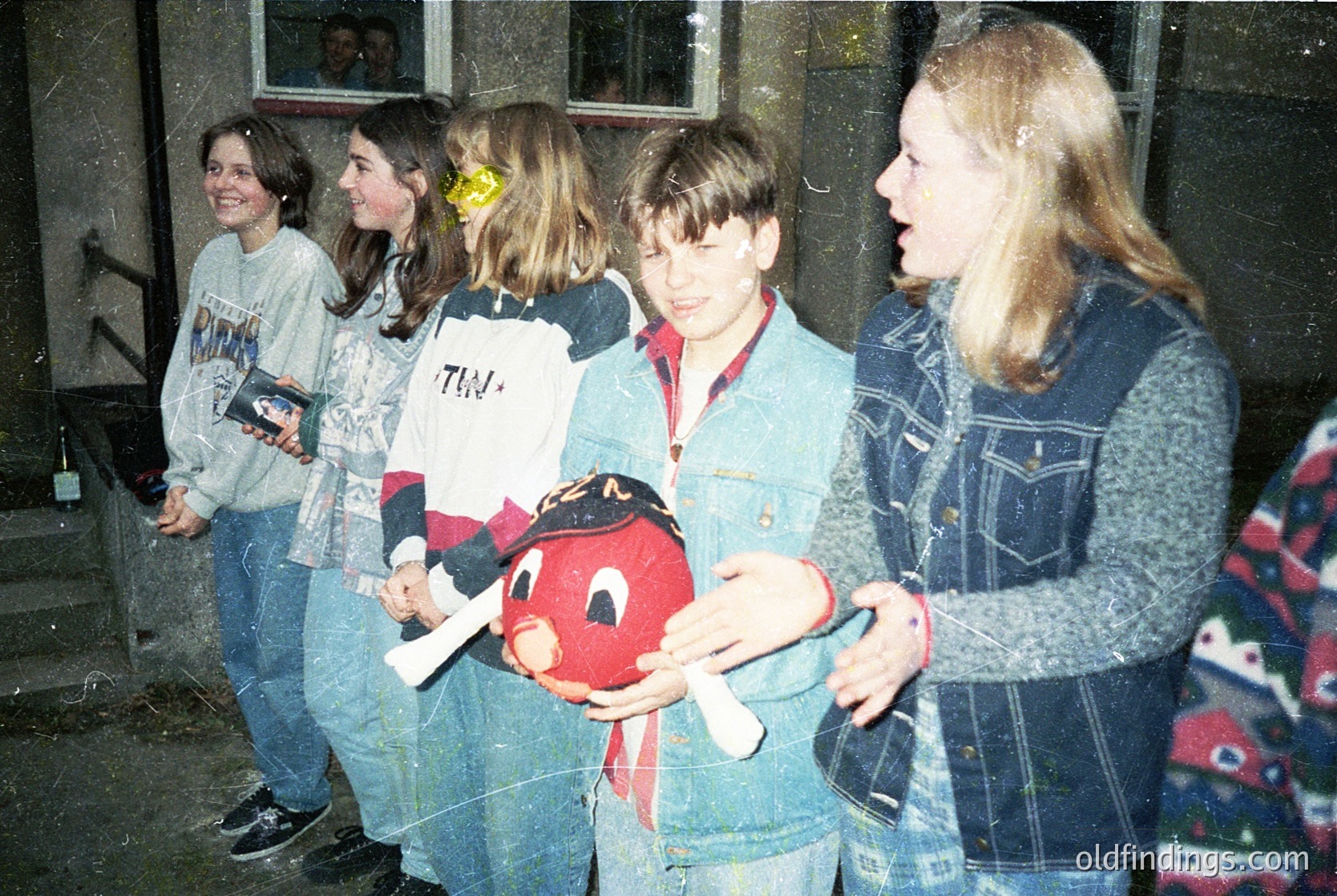 Vintage group photo featuring four teens in casual 1990s attire—flannel shirts, jeans, and a red plush toy (likely a *Troll* doll). Indoor setting with brick walls and a window. Youthful energy and playful expressions capture a moment of camaraderie.