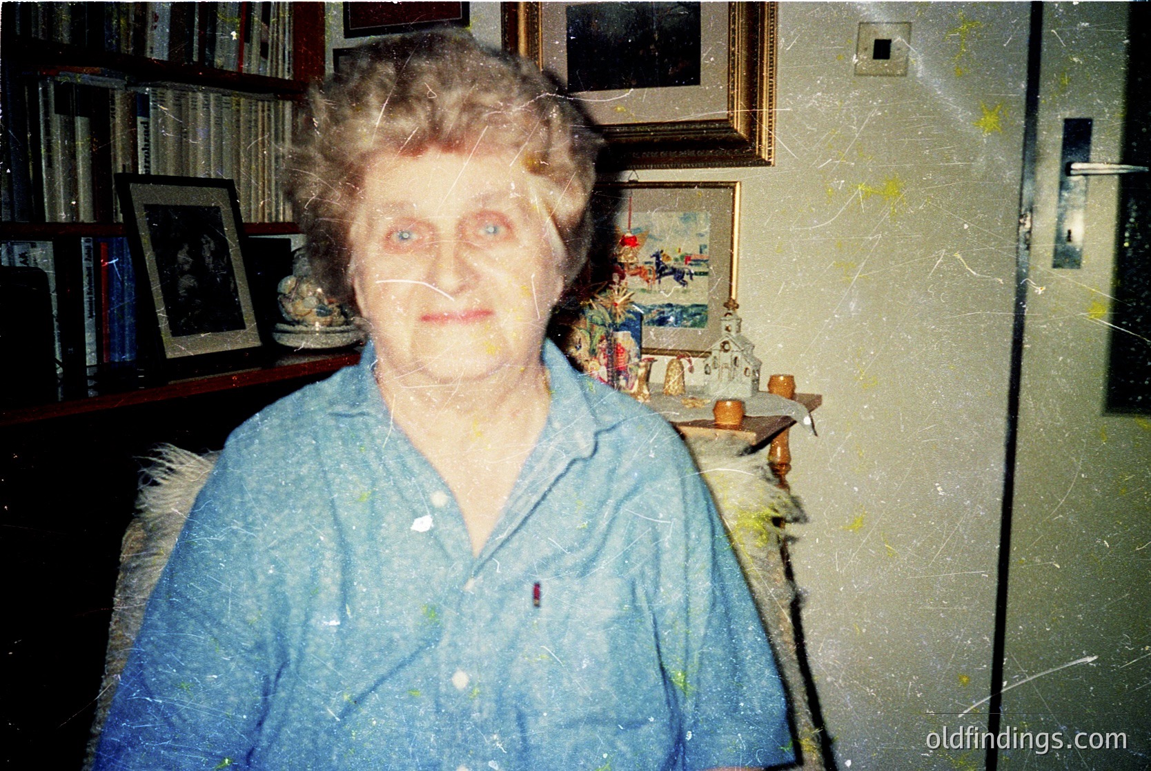 Portrait of an elderly woman in a vintage indoor setting, likely mid-20th century. She wears a light blue button-up shirt with a subtle patch on the left breast. Behind her, framed artworks, bookshelves, and a decorative shelf with small figurines and fruit create a lived-in, nostalgic atmosphere.