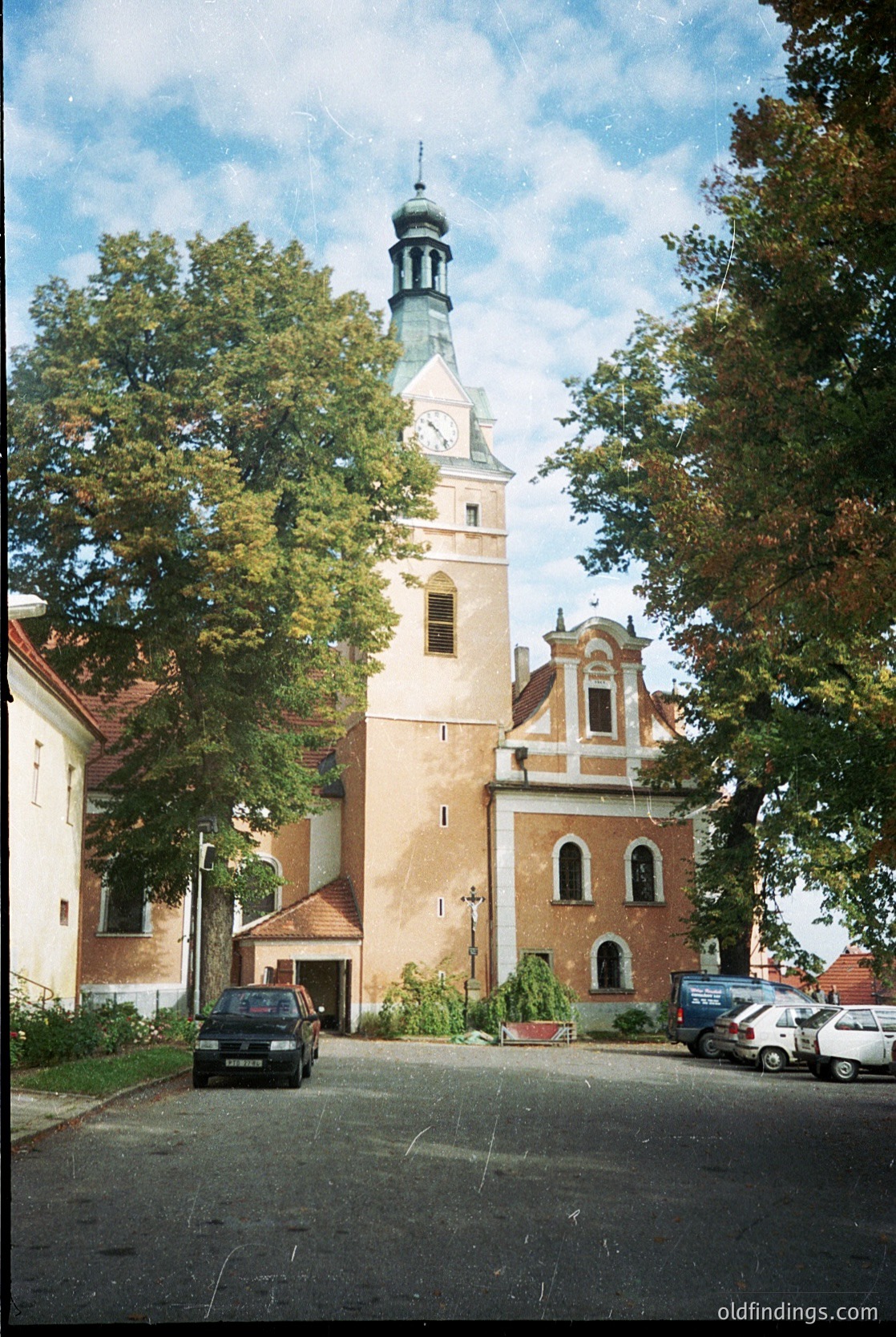 Historic church with Baroque-style tower and clock, featuring cream brickwork and arched windows. Surrounded by mature trees and parked cars indicating modern use. Likely Eastern European architecture, possibly .