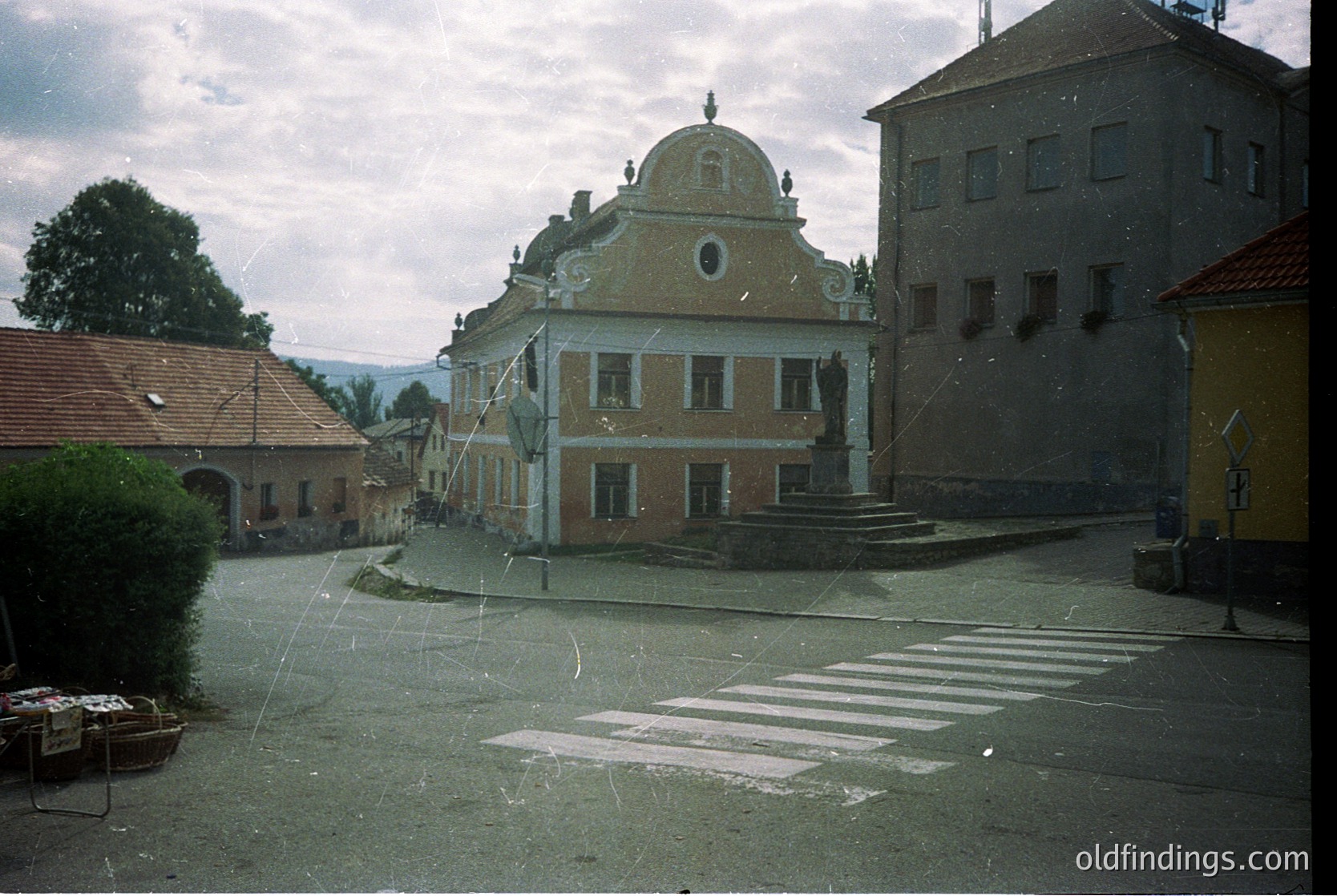Neoclassical building with domed roof and decorative facade in a European village square, likely 1970s–1980s. Crosswalk and vintage street signs suggest preserved urban design.