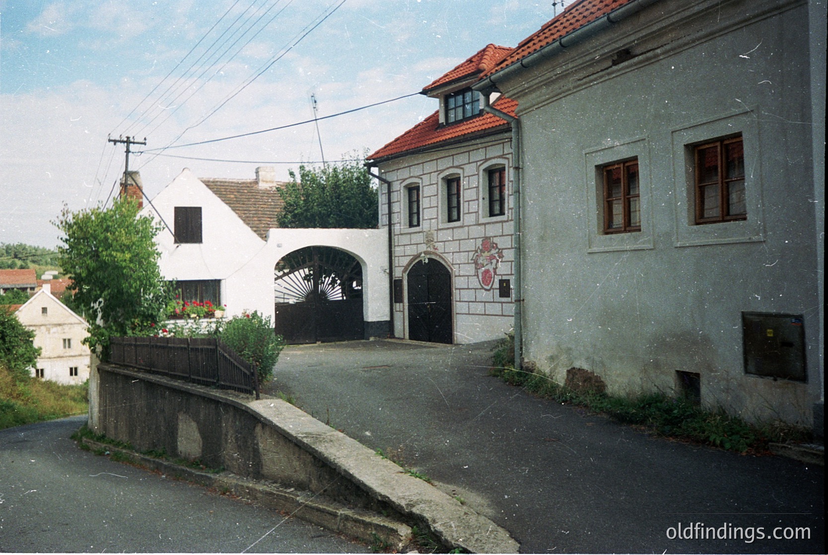 Eastern European village street with mid-20th century architecture—gray stucco buildings featuring arched entryways, wooden shutters, and red-tiled roofs. A small arched gateway leads to a courtyard, flanked by utility poles and overhead wires. Residential scale, likely 1960s-1980s.