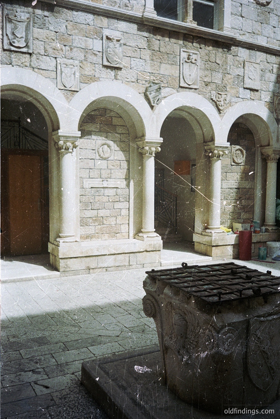 Neoclassical stone courtyard façade with three arched doorways, flanked by Corinthian columns and decorative reliefs. Stone drinking fountain with carved basin and spout in foreground. Weathered stonework suggests historic European architecture, likely 19th-century. Potential institutional or civic use.