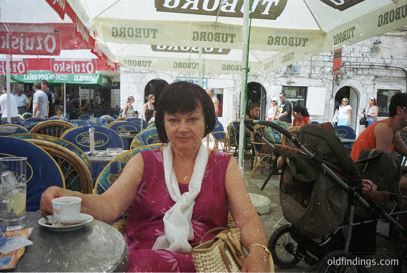 Vintage seaside café scene with woman in pink dress sipping coffee under a Tuborg beer umbrella. Stone-paved promenade, strollers, and retro seating in background. Likely Mediterranean coastal town, 1970s-1980s. é