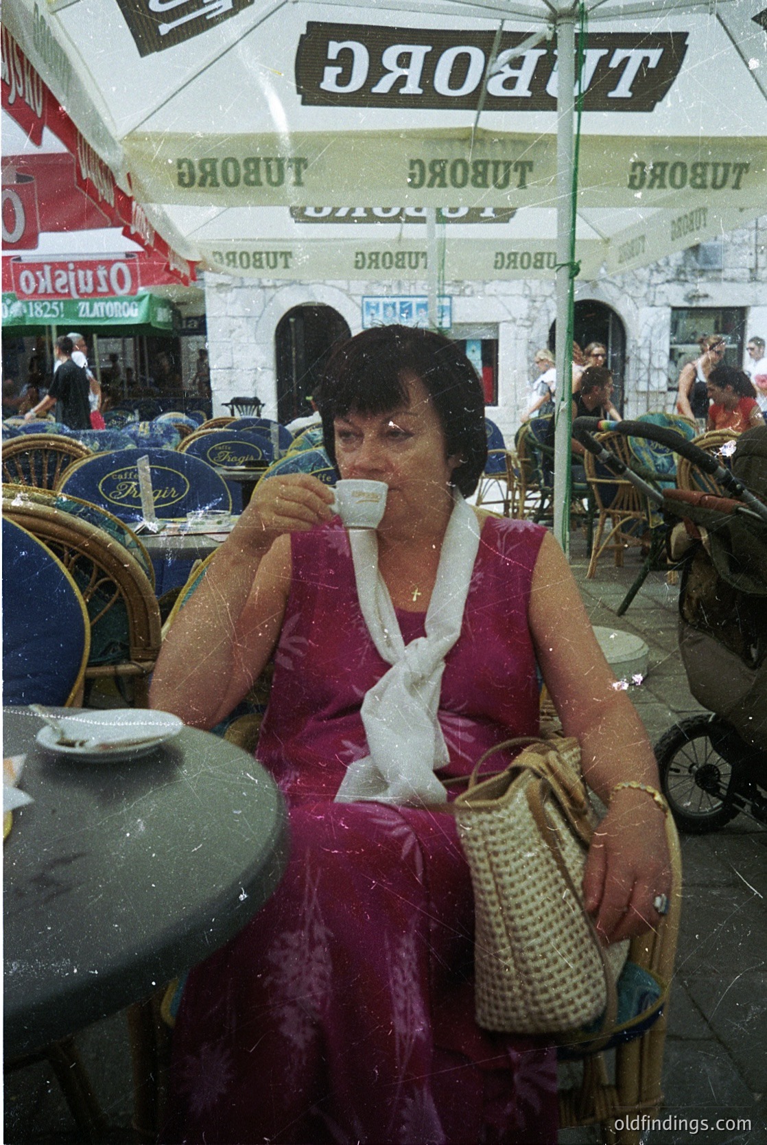 Mid-century café scene featuring a woman in a pink dress sipping coffee under a Tuborg-branded umbrella. Woven tote bag and white scarf add vintage charm. Outdoor seating with metal chairs and tables suggests a European seaside or beachside setting. Likely 1960s–1970s. é