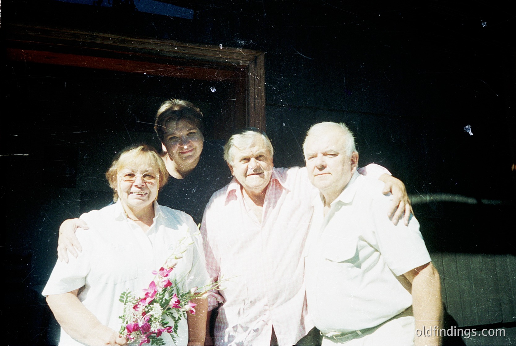 Four adults pose indoors, likely in a 1970s–1980s Eastern European setting. The woman holds pink flowers; men wear light-colored button-ups. Warm lighting and a wooden door frame suggest a domestic or celebratory occasion.