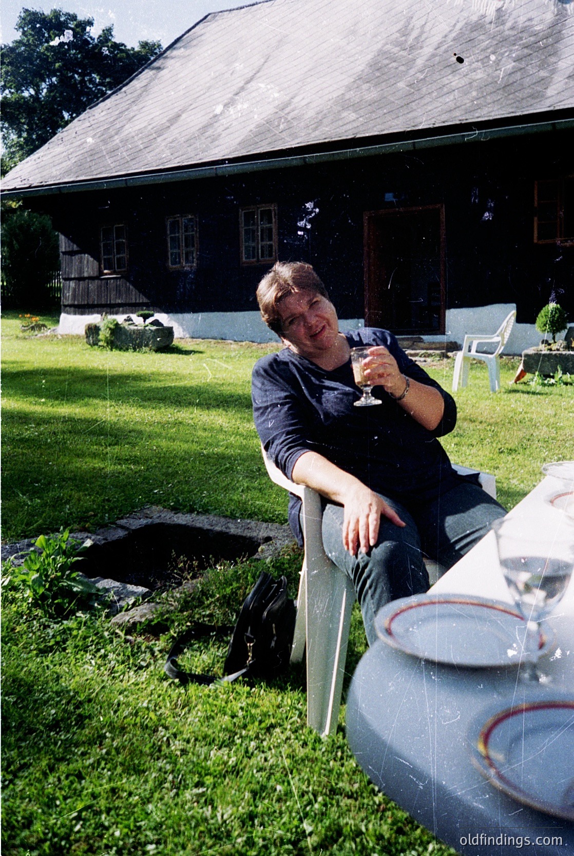 Relaxed individual in outdoor setting, holding a wineglass beside a rustic wooden house with white trim. Mid-century style furniture (white plastic chair) and vintage metal urn on grassy lawn. Lush greenery and mature trees in background.