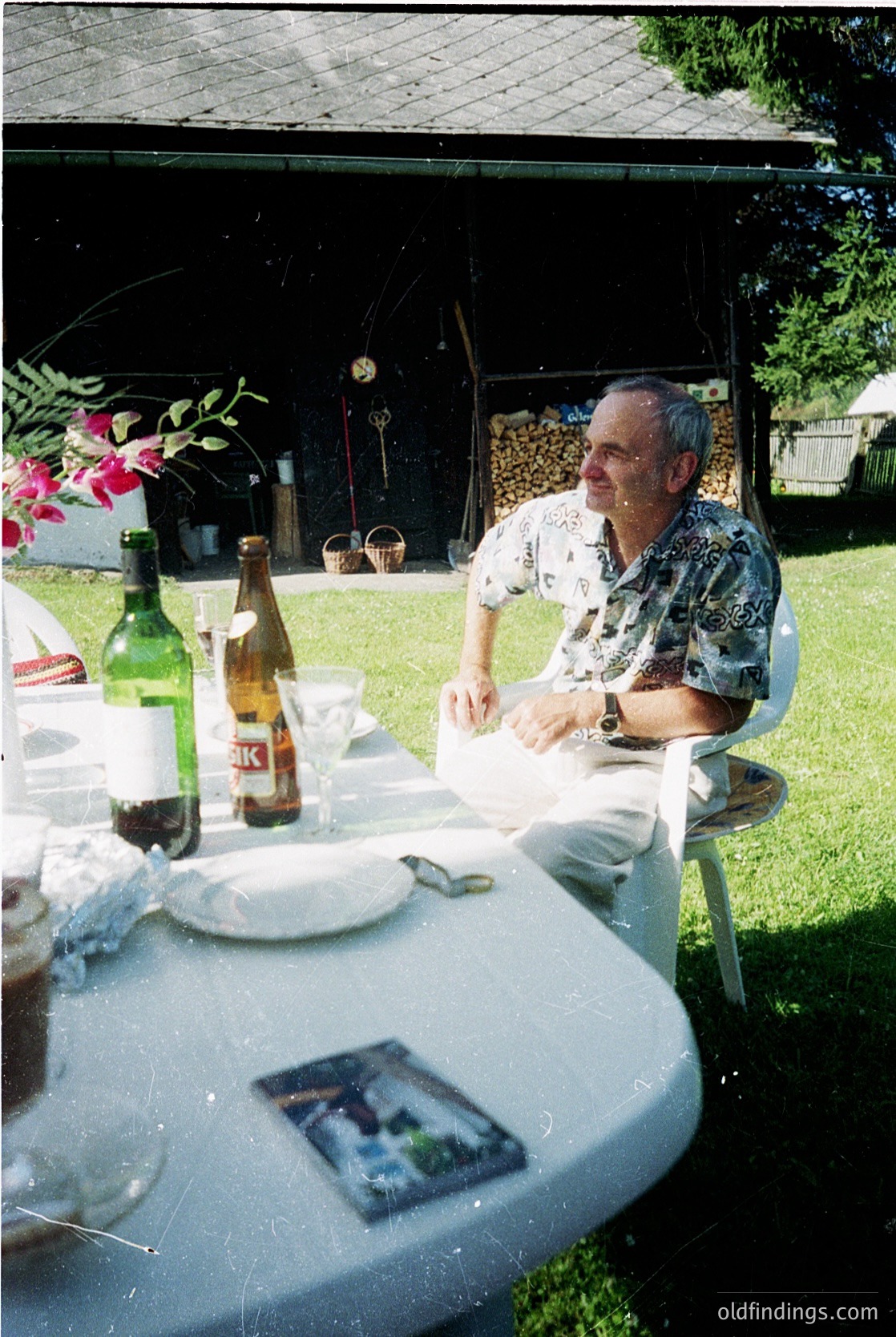 Vintage outdoor dining scene featuring a man in a floral-patterned shirt seated at a white-clothed table under a shaded canopy. Bottles of beer (Kastel brand) and a wine bottle with a white label sit beside a napkin and small photo. Lush greenery and a woodpile in the background suggest a garden setting, likely mid-20th century.