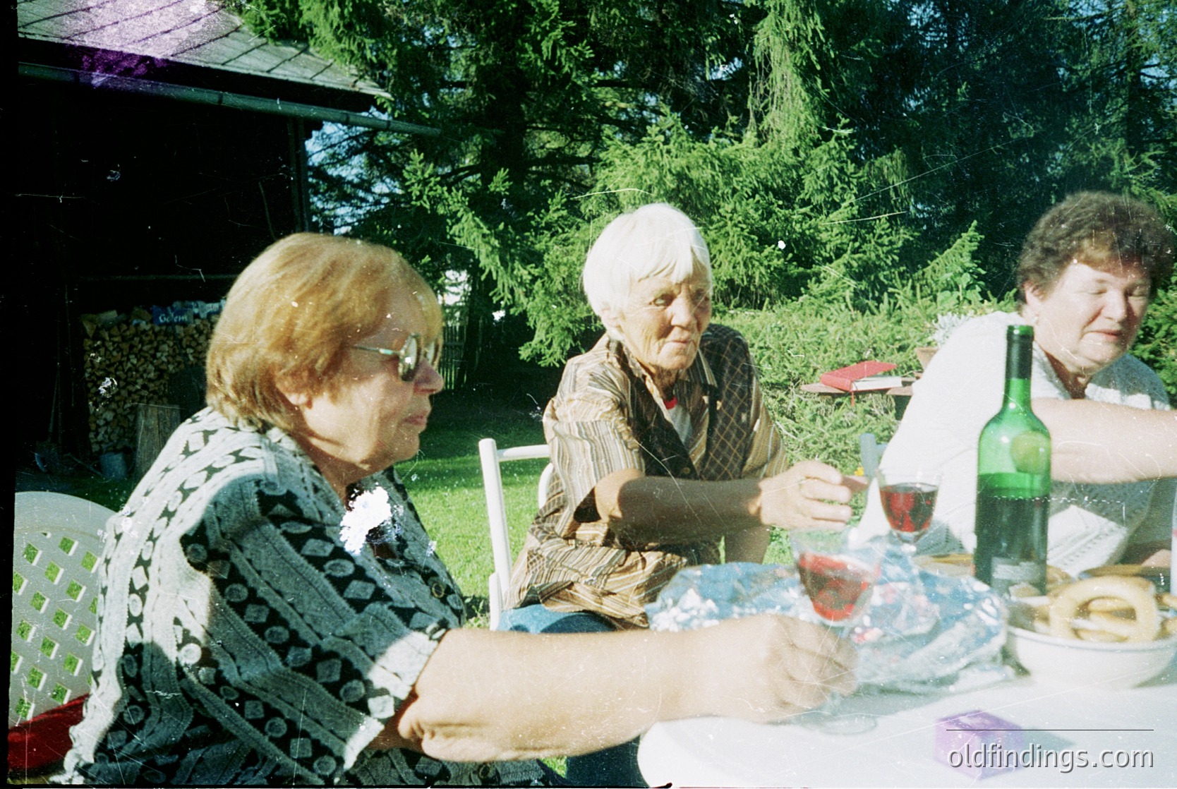 Three elderly women seated outdoors at a picnic-style table, enjoying wine and conversation. Mid-20th century clothing (1970s-1980s) and vintage camera grain suggest a personal or family gathering. Lush greenery and wooden structure hint at a rural or suburban backyard setting.