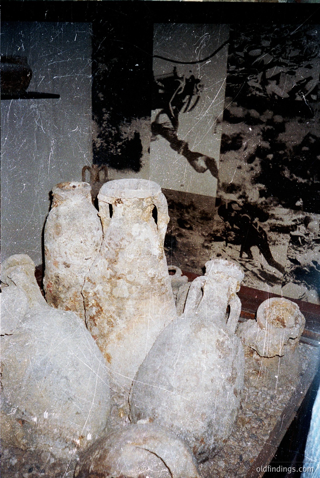 Ancient Greek pottery fragments, including two intact amphorae and broken vessels, displayed in a dimly lit archaeological setting. Surface erosion and mineral deposits suggest long-term exposure. Likely from a 5th–4th century BCE Greek site.