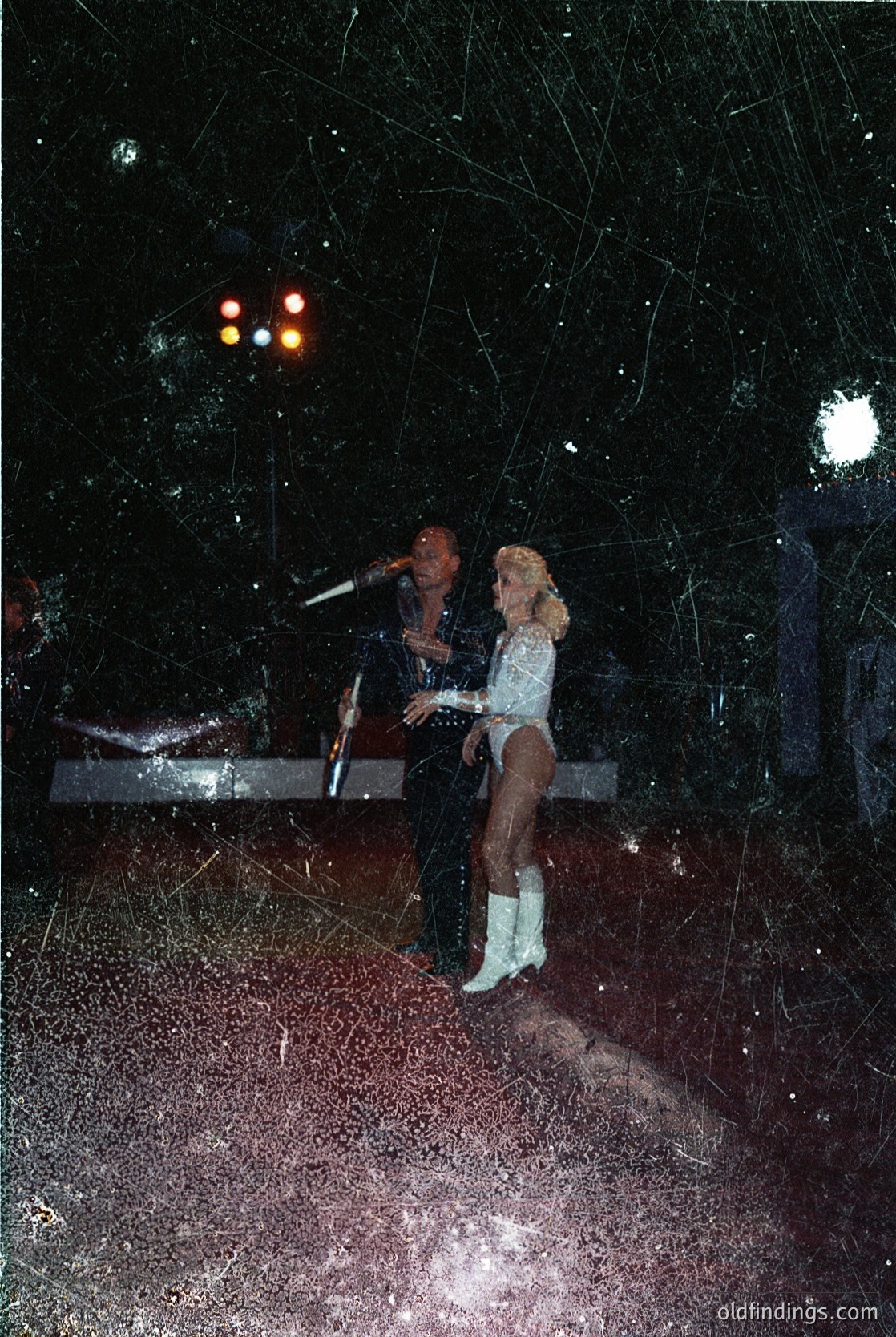 A low-angle nighttime shot of a couple dancing in a dimly lit, possibly outdoor venue. The woman wears a sleeveless white dress with a cowboy-style belt and boots, while the man holds a microphone, suggesting live entertainment. Decorative lights and strings create a festive atmosphere. Style hints at 1970s-1980s Western or country music culture.