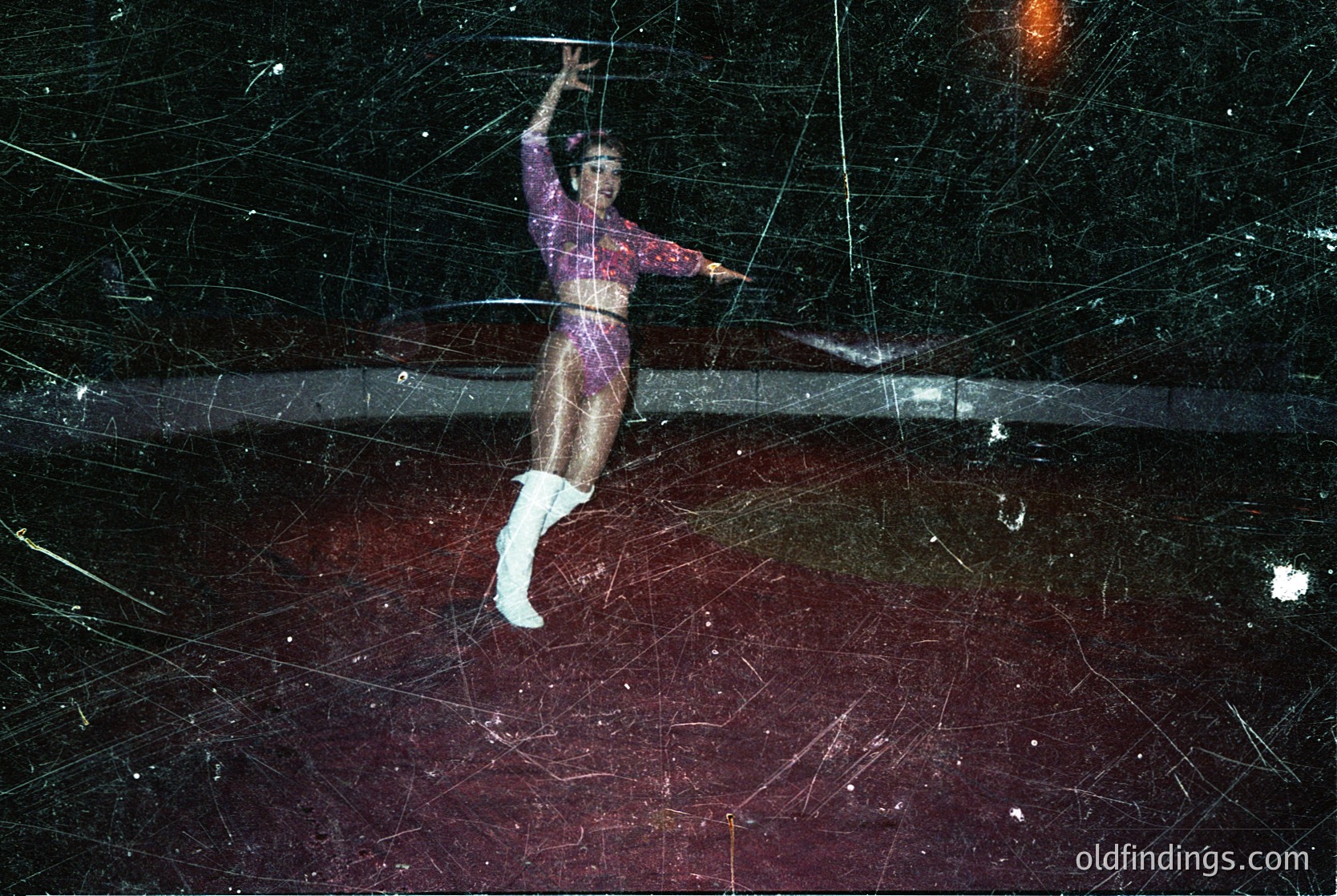 Circus performer mid-air on trapeze bar, dressed in 1970s-style leotard with geometric patterns and white boots. Dynamic shot captures motion blur of stage lights and audience below. High-energy performance art, likely from a vintage circus or variety show.