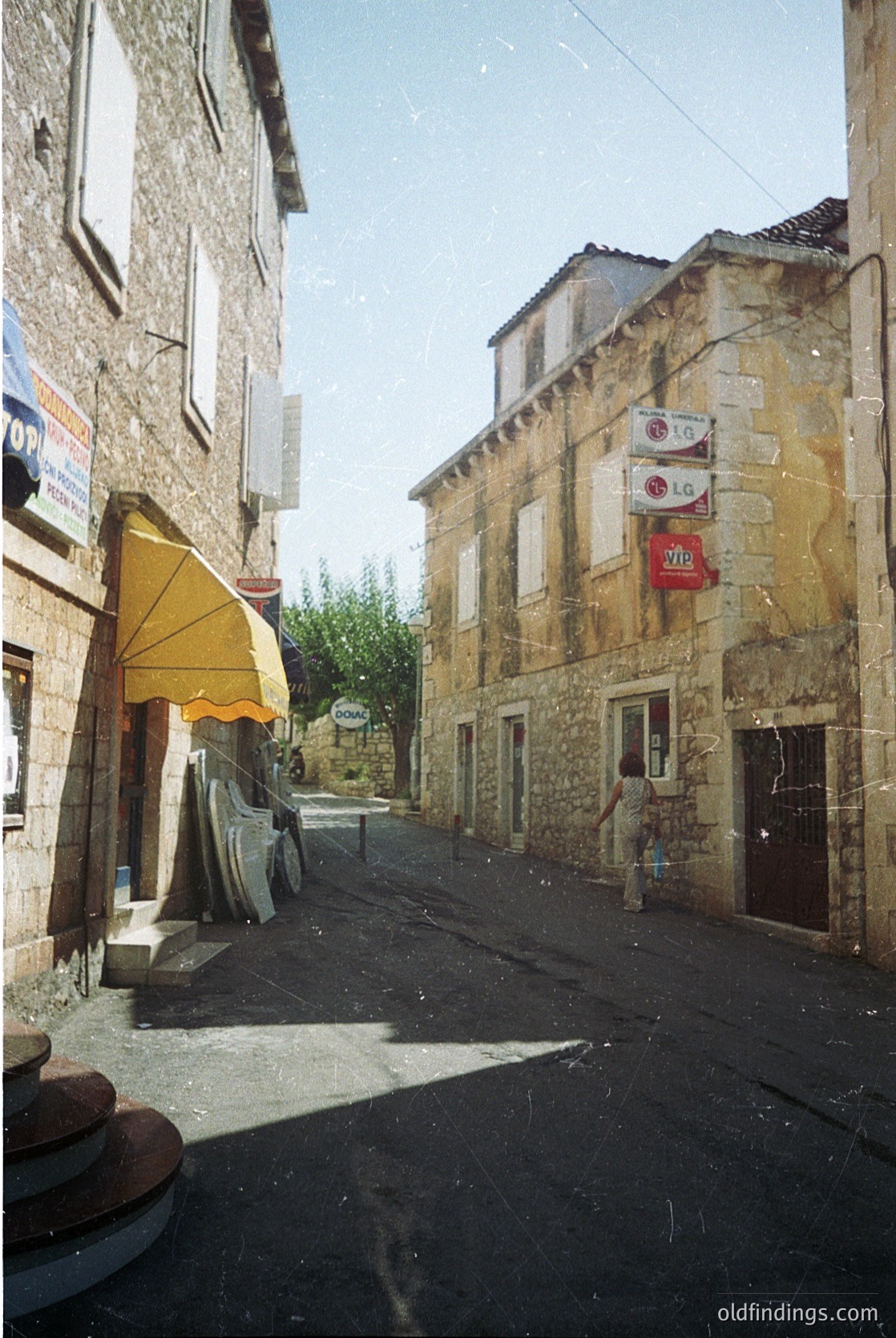 Stone-built European village street with rustic charm, featuring weathered facades and shuttered windows. A yellow awning extends over a doorway, while red signs indicate local businesses. Cobblestone path and vintage signage suggest historic preservation. Likely , France, -2000s.
