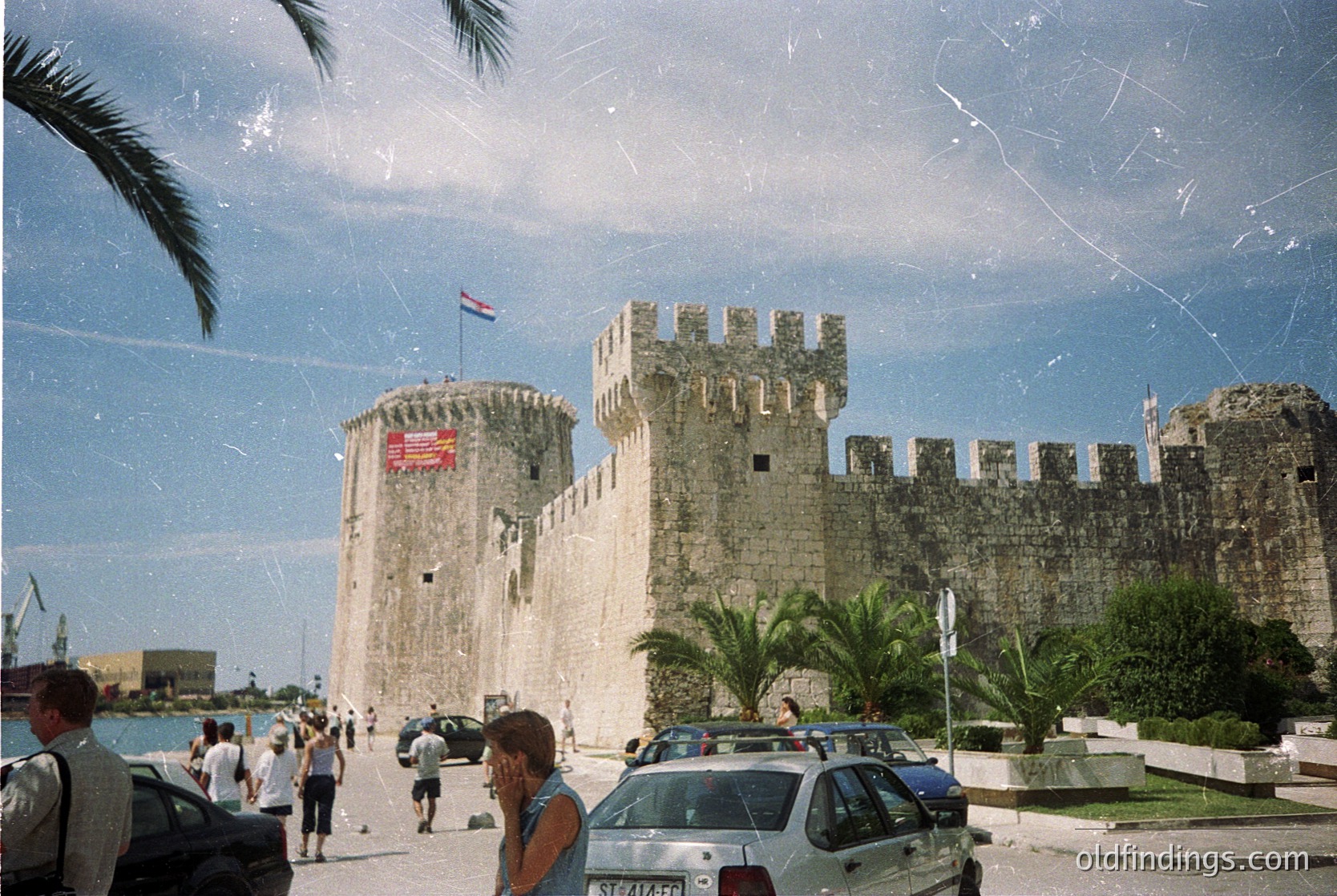 Medieval fortress walls with crenellations line a seaside promenade in Split, Croatia. A prominent tower displays a Croatian flag atop, with a US flag banner mid-construction. Palm trees and modern cars contrast with historic architecture. Crowds stroll along the waterfront, suggesting a popular tourist destination.