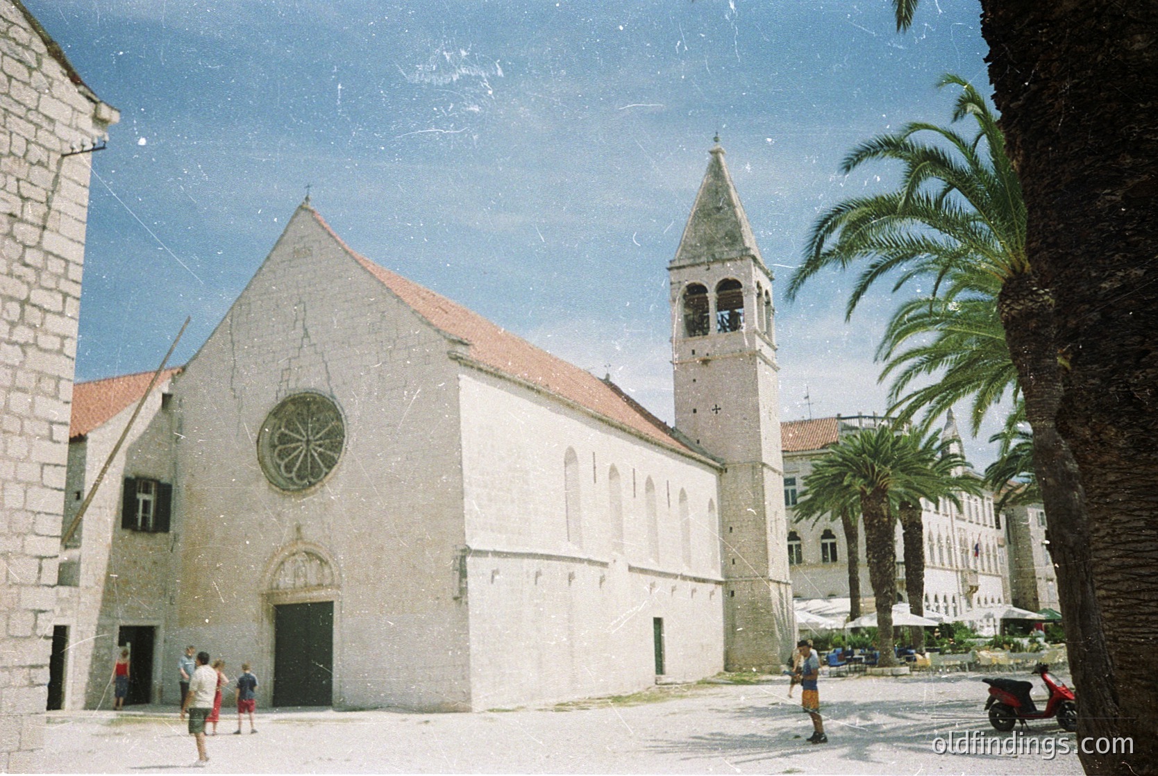 Medieval stone church with round rose window and bell tower, set in a Mediterranean courtyard. Light-colored facade contrasts with terracotta roof. Palm trees and modern pedestrians suggest coastal urban setting. Likely –15th century European architecture. #