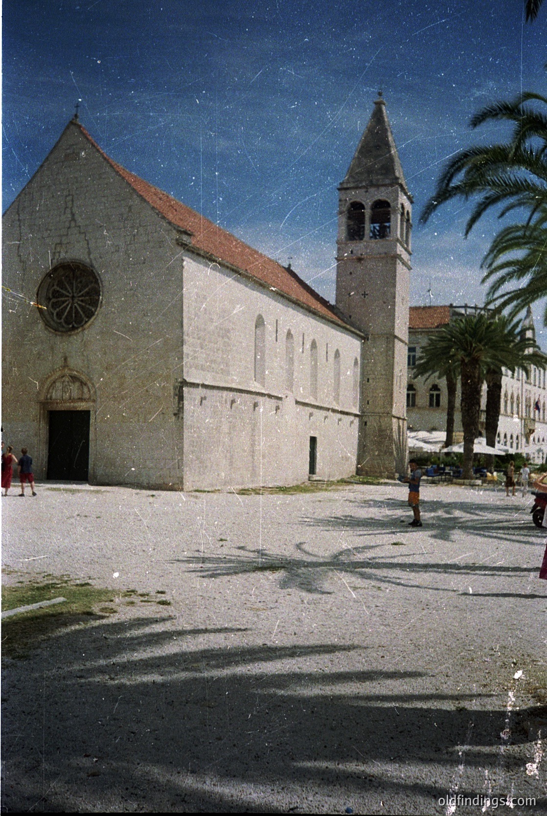 Romanesque-style church with round-arched doorway and rose window, set on a cobblestone plaza. Tall bell tower with a pointed roof and small openings. Palm trees and Mediterranean architecture in background. Likely coastal European location, possibly --- *Note: The architectural style and cobblestone plaza suggest a Mediterranean coastal city, with Dubrovnik (Croatia) as a strong possibility due to its well-preserved Romanesque/Renaissance buildings. The palm trees and overall aesthetic align with the region’s historic charm.*