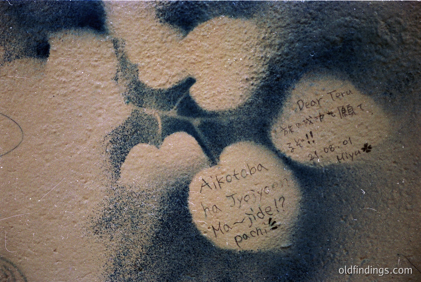 Graffiti-style handwritten messages in Japanese on a concrete surface, likely a sidewalk or plaza. Visible text includes "Aikotoba ha Jyuryo? Ma-yide!?" and "Dear Teru 21.08.01 Hiyu." Date suggests early 2000s. Urban street art reference with cultural/linguistic focus.