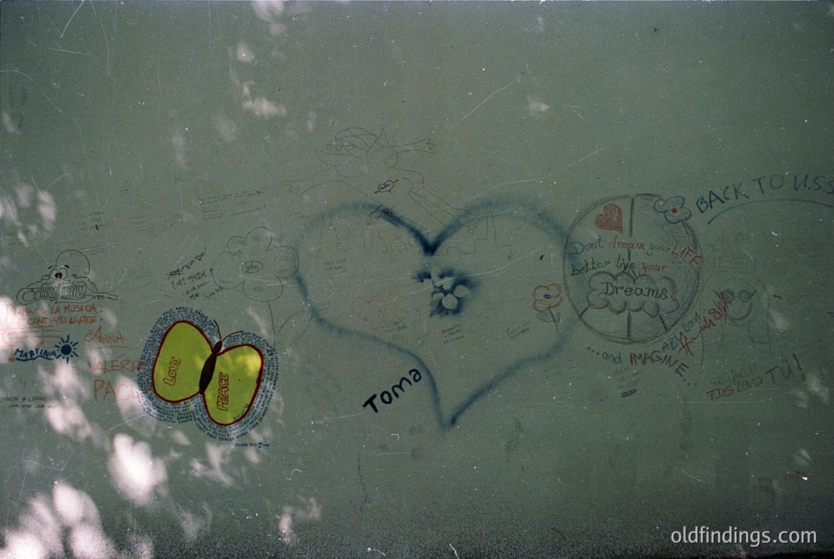 Graffiti-covered glass surface featuring handwritten messages, including a large heart with "Toma" inscribed, a yellow heart symbol, and a circular quote: *"Back to U... Don’t dream your life, imagine yours."* Likely a public or historical site. *(Note: Exact location/time uncertain; context inferred from style and content.)*