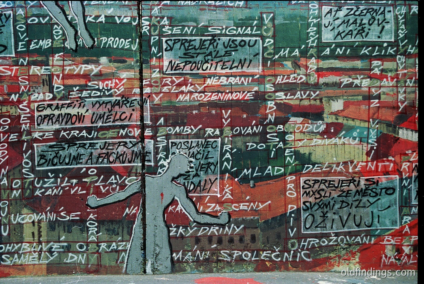 Graffiti-style mural with layered Czech/Slovak text on weathered concrete, featuring bold red and white paint. Central figure appears to be a stylized hand or arm holding a sign. Text includes warnings, slogans, and political/activist messages. Likely from the 1980s–1990s Eastern Bloc era, possibly Prague or Brno.