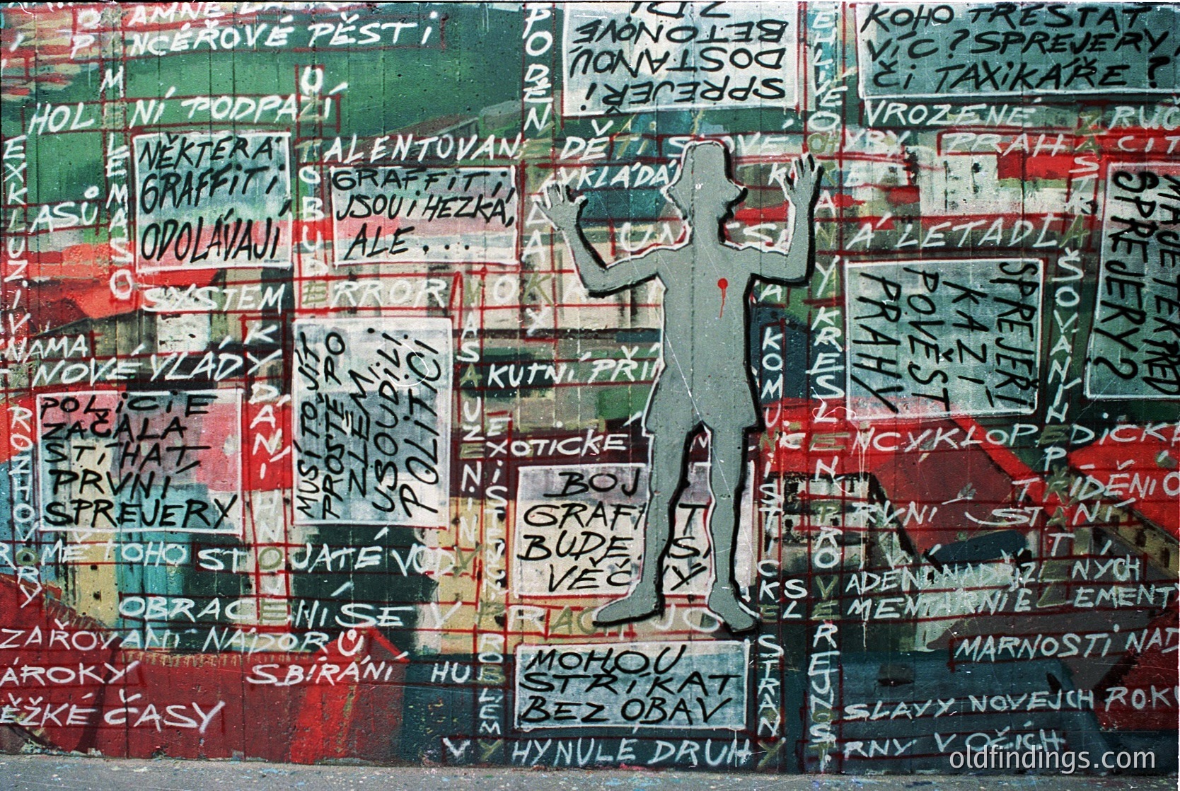 Vibrant graffiti mural featuring bold, layered text in Cyrillic script with a mix of red, green, and white colors. Central silhouette of a figure in mid-jump against a red background. Appears to be a political/social commentary piece, likely from Eastern Europe. Collage-style with overlapping signs and slogans.