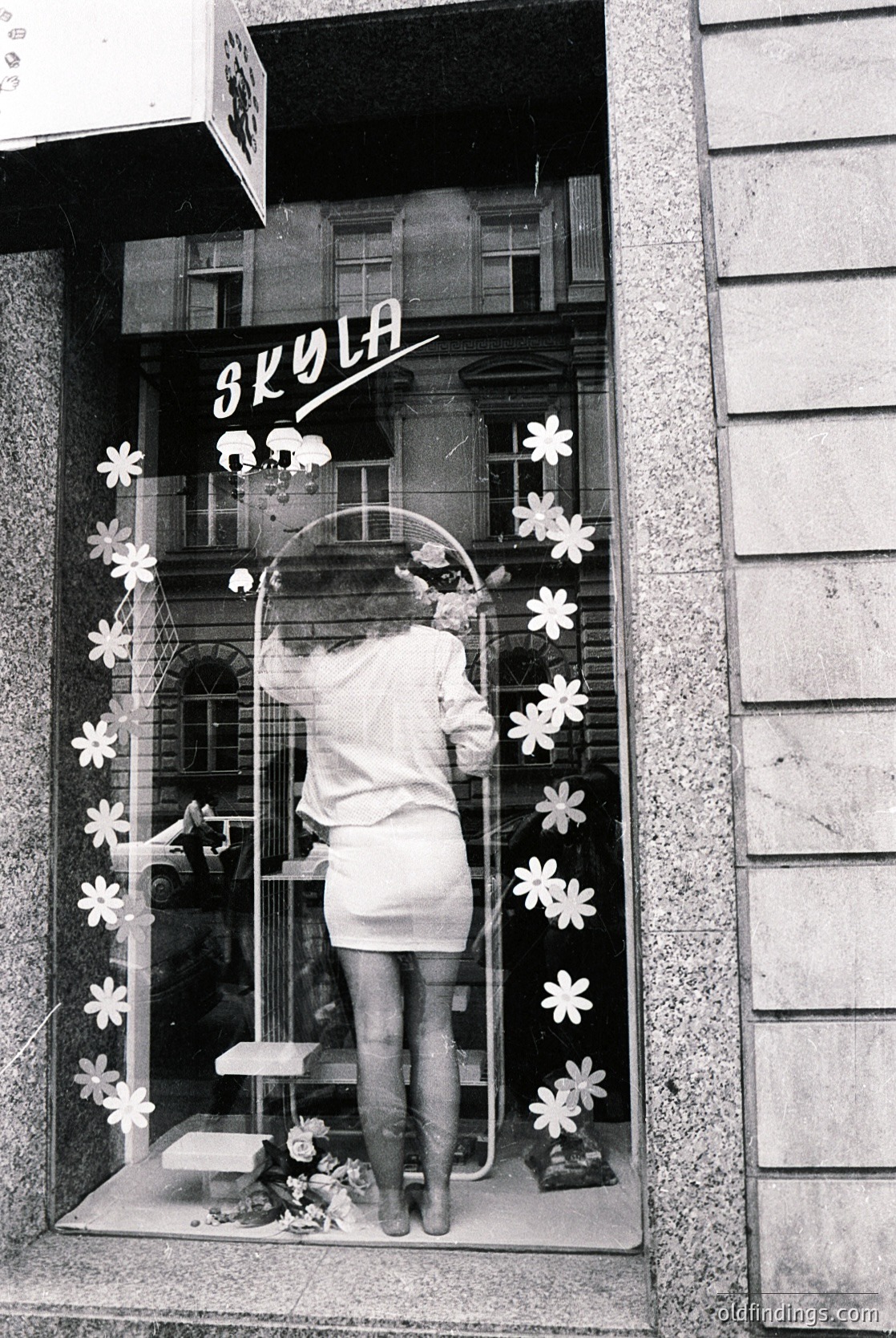 Mid-century school display featuring floral window art and Cyrillic sign "СКОЛА" (Bulgarian for "school"). Reflective glass captures a child’s silhouette in a short dress, standing on a small platform. Decor includes paper flowers, a circular frame, and a vase with dried flowers. Likely Eastern Bloc-era educational institution, 1960s–1980s.