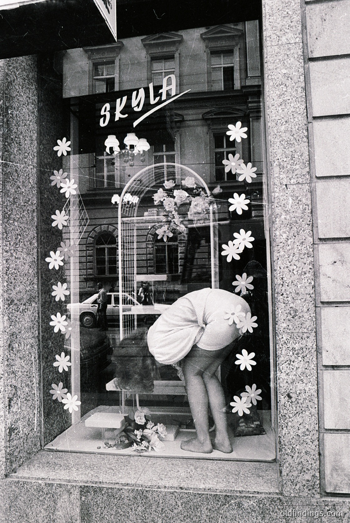 Retro storefront display for "Skyla" featuring floral decals and a mannequin in a white dress. Reflective window reveals vintage car and urban street scene. Likely 1960s–1970s European fashion retail.