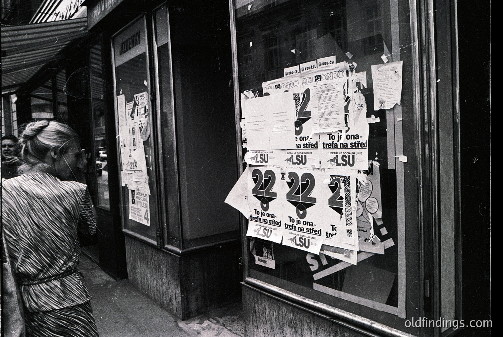 Black-and-white street scene featuring a woman in a striped dress examining posters taped to a storefront window. Prominent "22" price signs and text in Polish ("To ja ona-trefa na stref") suggest a 1970s–80s Polish market or collective shop. Urban, candid, and culturally specific.
