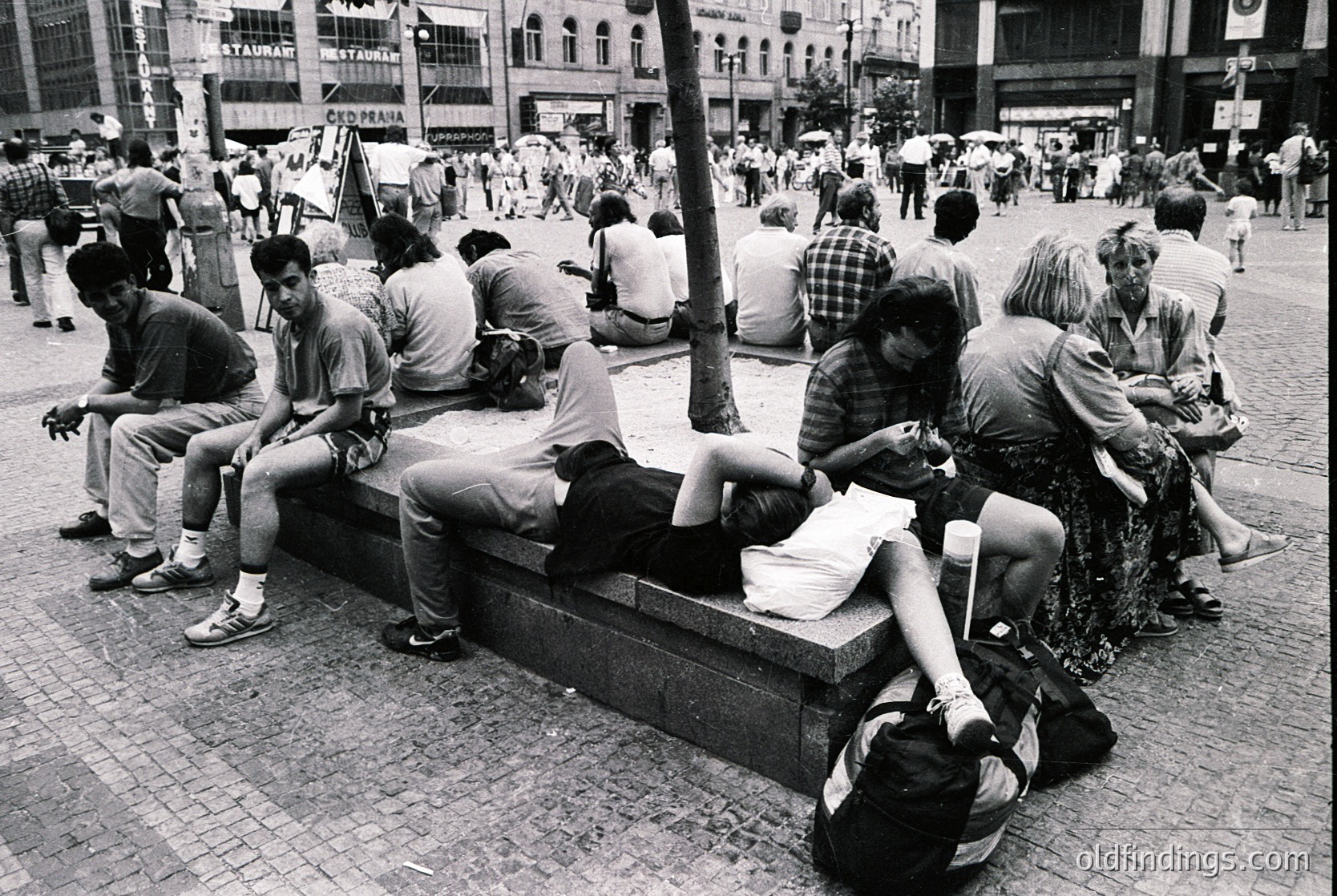 Street scene from the 1980s–1990s featuring a diverse group of people relaxing on a stone bench in a busy urban plaza. Casual attire includes jeans, t-shirts, and backpacks; one person holds a rolled poster. Surrounding buildings display commercial signage in Cyrillic script, suggesting Eastern Europe. Crowds fill the background, indicating a public gathering spot.