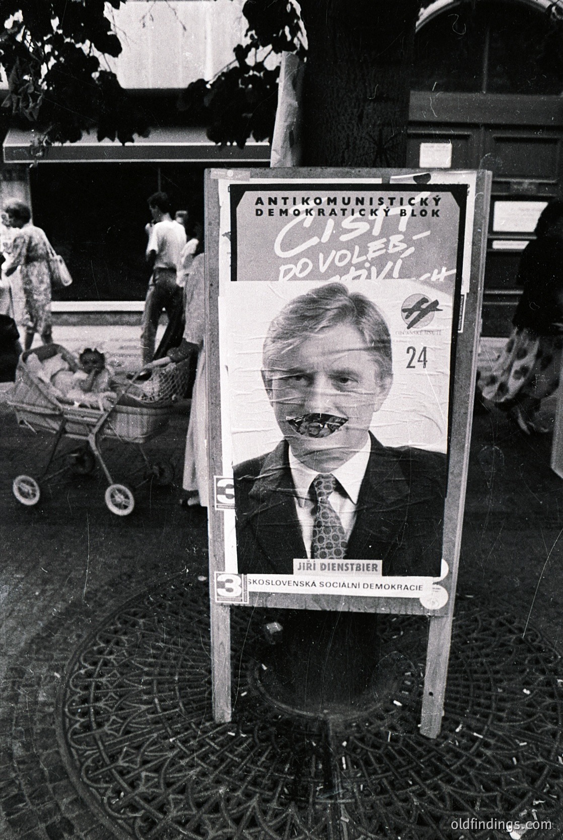 Vintage political campaign poster featuring Jiří Dienstbier, leader of the *Antikomunistický demokratický blok* ("Anti-Communist Democratic Bloc") in Czechoslovakia, advocating for democratic elections. The poster, dated 1990, includes a caricatured face with exaggerated teeth, emphasizing anti-communist sentiment. Urban outdoor setting with blurred pedestrians and a child in a stroller. --- *Note: "Jiří Dienstbier" and "Czechoslovakia" are used for specificity; "ColdWarPolitics" and "DemocraticTransition" enhance historical research value.*