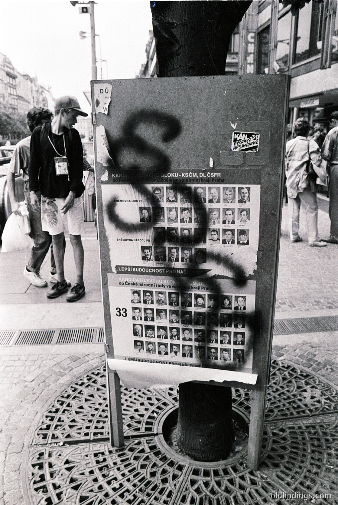 Vintage public notice board in urban setting, covered in graffiti (symbol resembling "S" or "Z"). Grid of black-and-white photos (likely missing persons or wanted posters) with Czech text. Street scene with pedestrians in casual summer attire. Metal grate and cobblestone pavement visible.