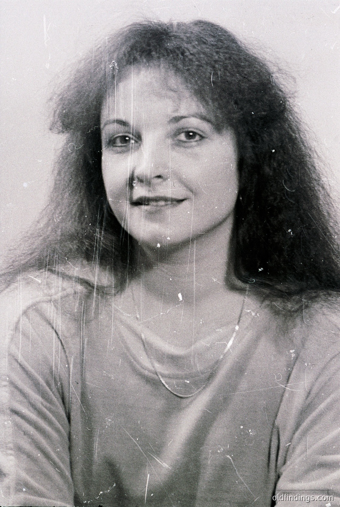 Portrait of a woman with voluminous, shoulder-length curly hair, shot in black-and-white. She wears a simple necklace and a light-colored blouse. The image appears aged with visible scratches and slight grain, suggesting a vintage or style. Her direct gaze and neutral expression convey timeless elegance.