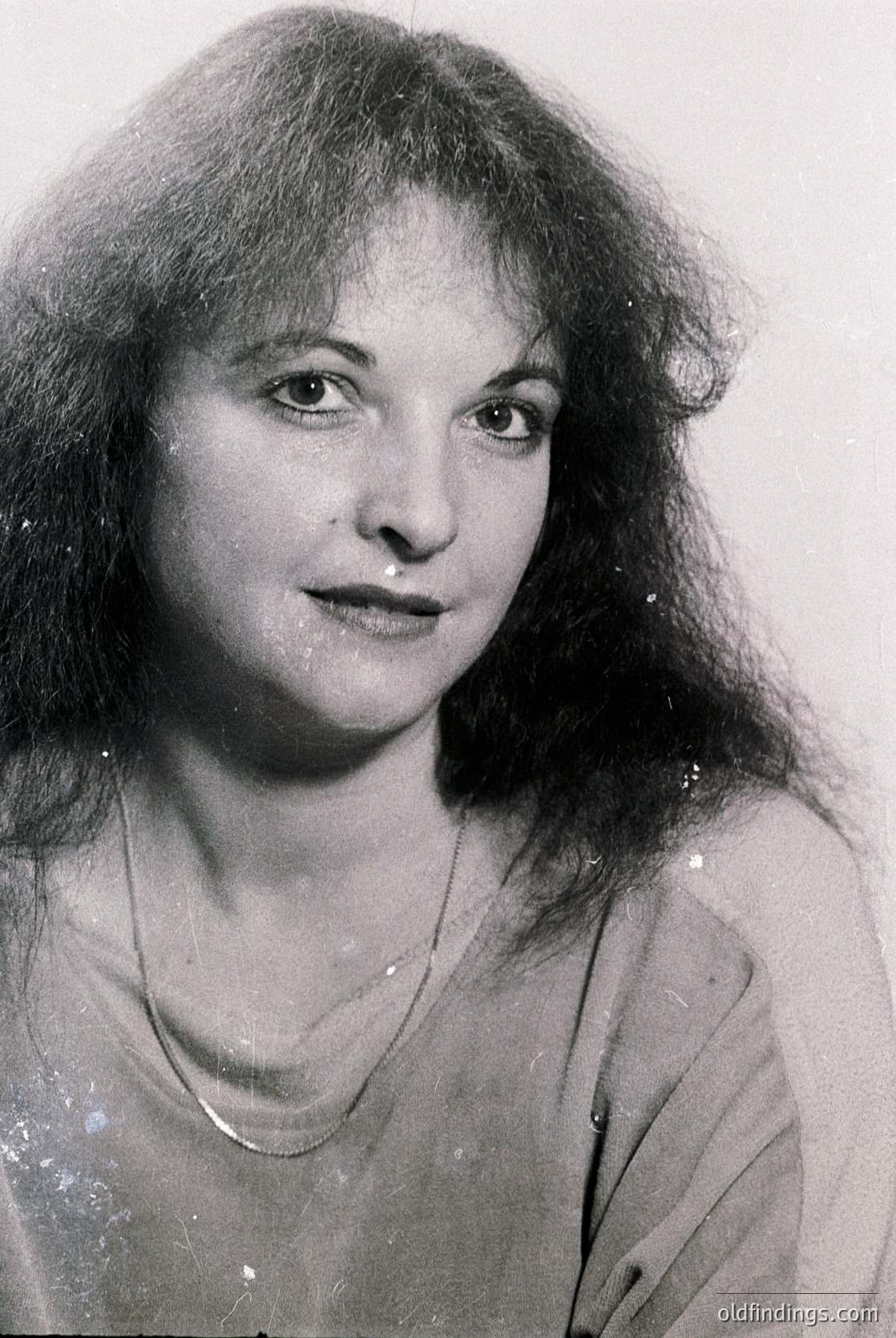 Portrait of a woman with voluminous shoulder-length wavy hair, shot in black-and-white. She wears a simple necklace and a loose blouse with a subtle V-neck. The lighting highlights her expressive face, suggesting a candid or documentary-style photograph. Likely from the mid-20th century (1950s–1970s).
