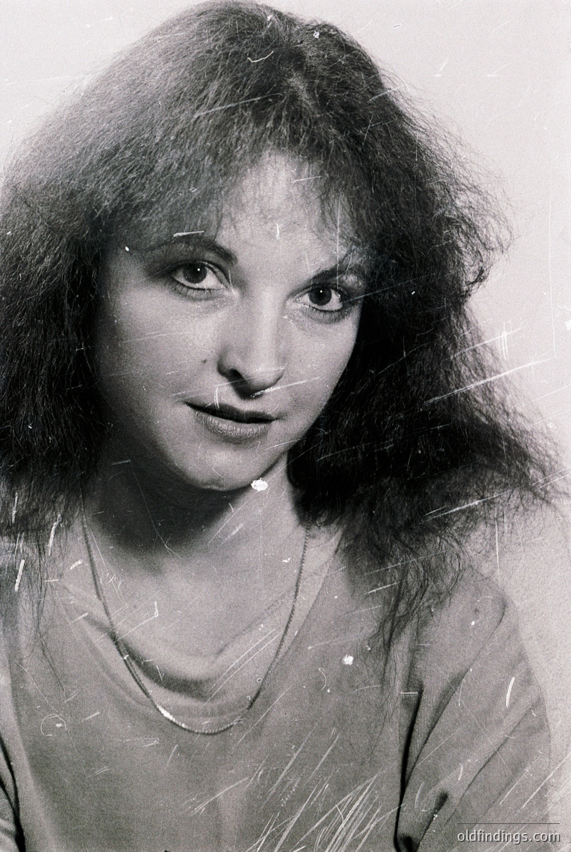 Portrait of a woman with voluminous shoulder-length hair, shot in black-and-white. Her expression is neutral, with a slight upward gaze. She wears a simple necklace and a high-necked top. Style suggests 1970s–1980s fashion.