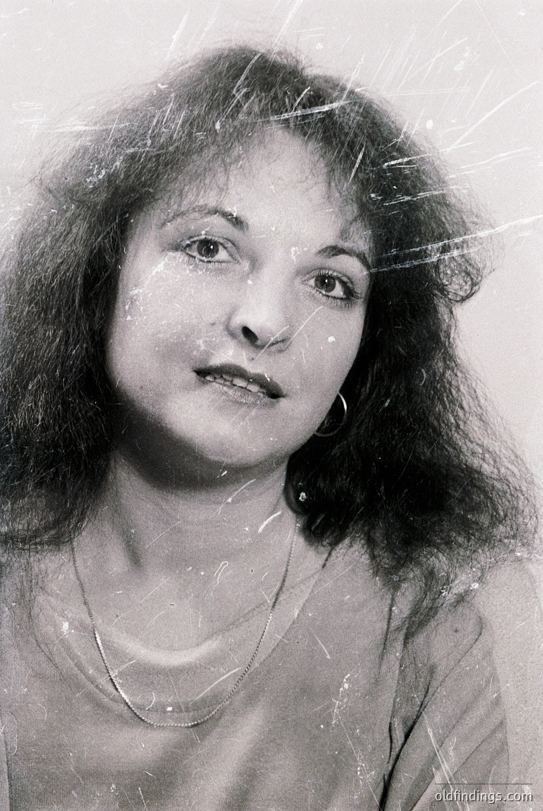 Portrait of a woman with voluminous shoulder-length hair, captured in monochrome. Snowflakes lightly dust her face and shoulders, suggesting winter or cold weather. Minimalist jewelry—a delicate necklace—complements her expression. Style evokes 1960s–1970s fashion.