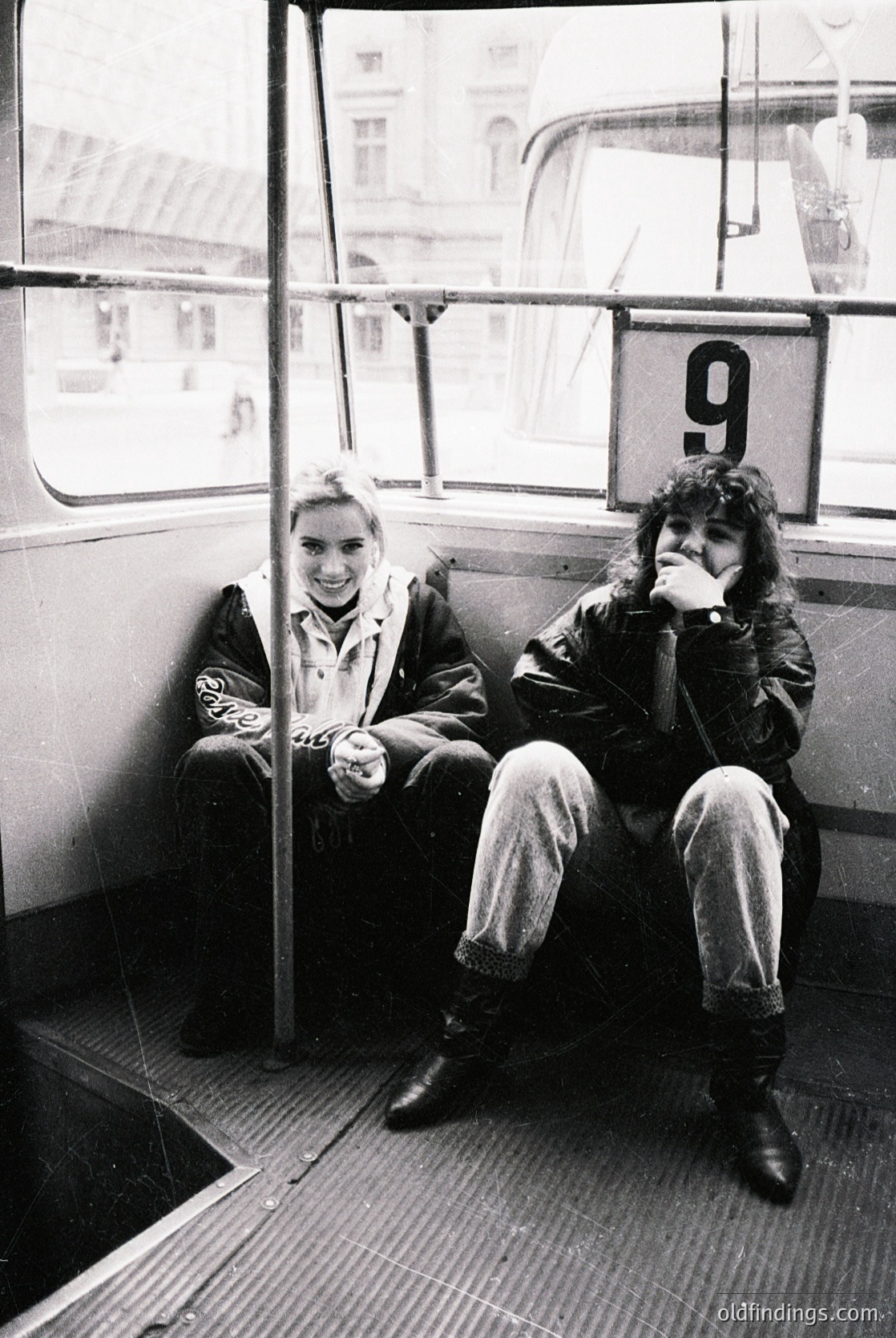 Two individuals seated in a vintage tram marked with the number **9**, likely from a European city. The man on the left wears a patterned sweater and light-colored pants; the woman on the right has curly hair and a dark jacket. Urban architecture and tram tracks visible outside. Style suggests **1970s–1980s** public transport.