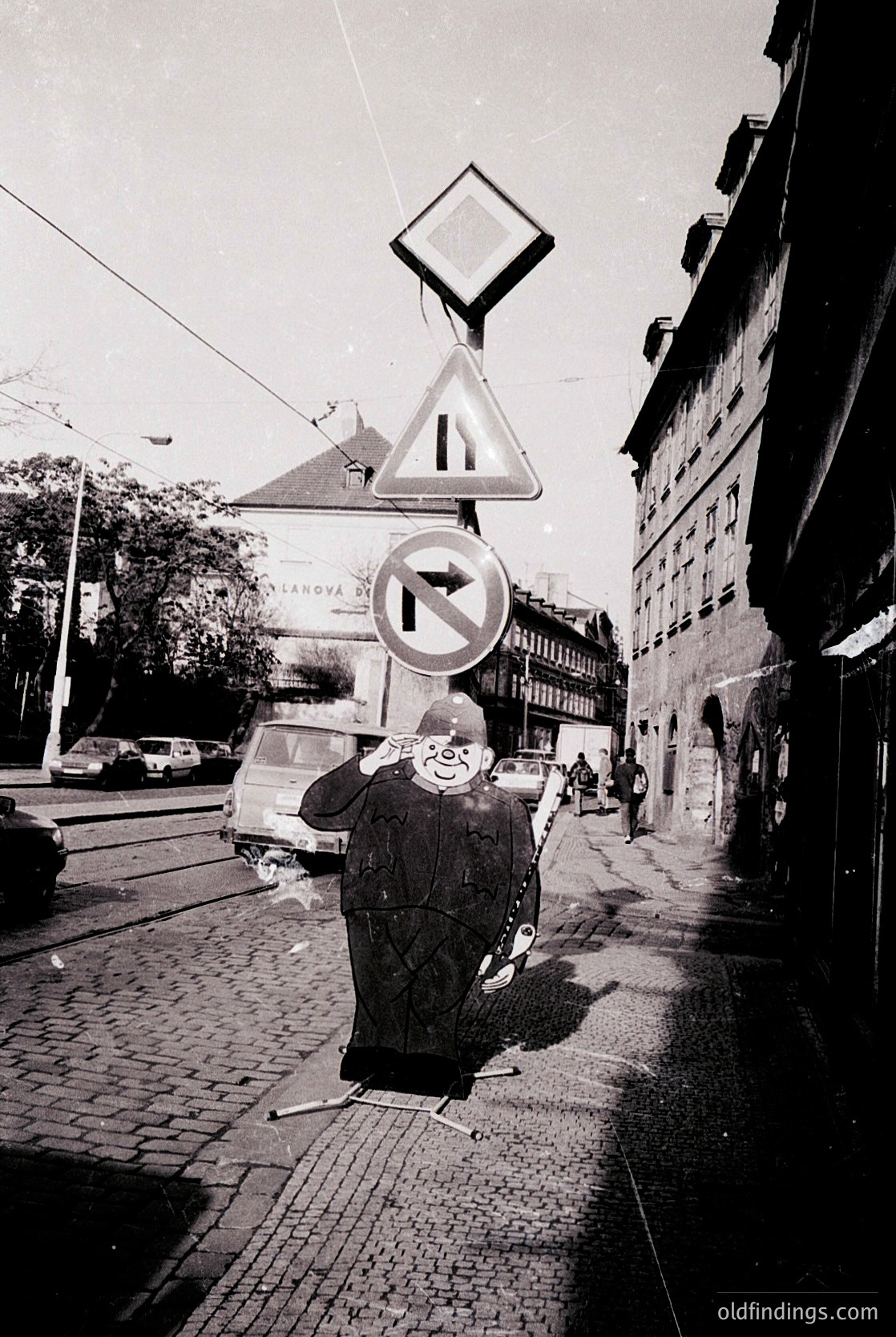 Mid-20th century urban street scene featuring a man in a long coat and hat crossing a cobblestone roadway. Traffic signs (yield and no entry) and tram wires indicate European city infrastructure. Buildings exhibit classic European architecture with brick facades and large windows. Likely --- *Note: The specific location appears to be Prague, Czech Republic, based on architectural cues and signage.*